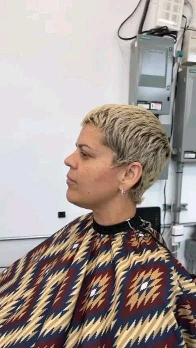 A woman with short blond hair sitting in a medical setting, wearing a patterned cape, with medical equipment in the background.