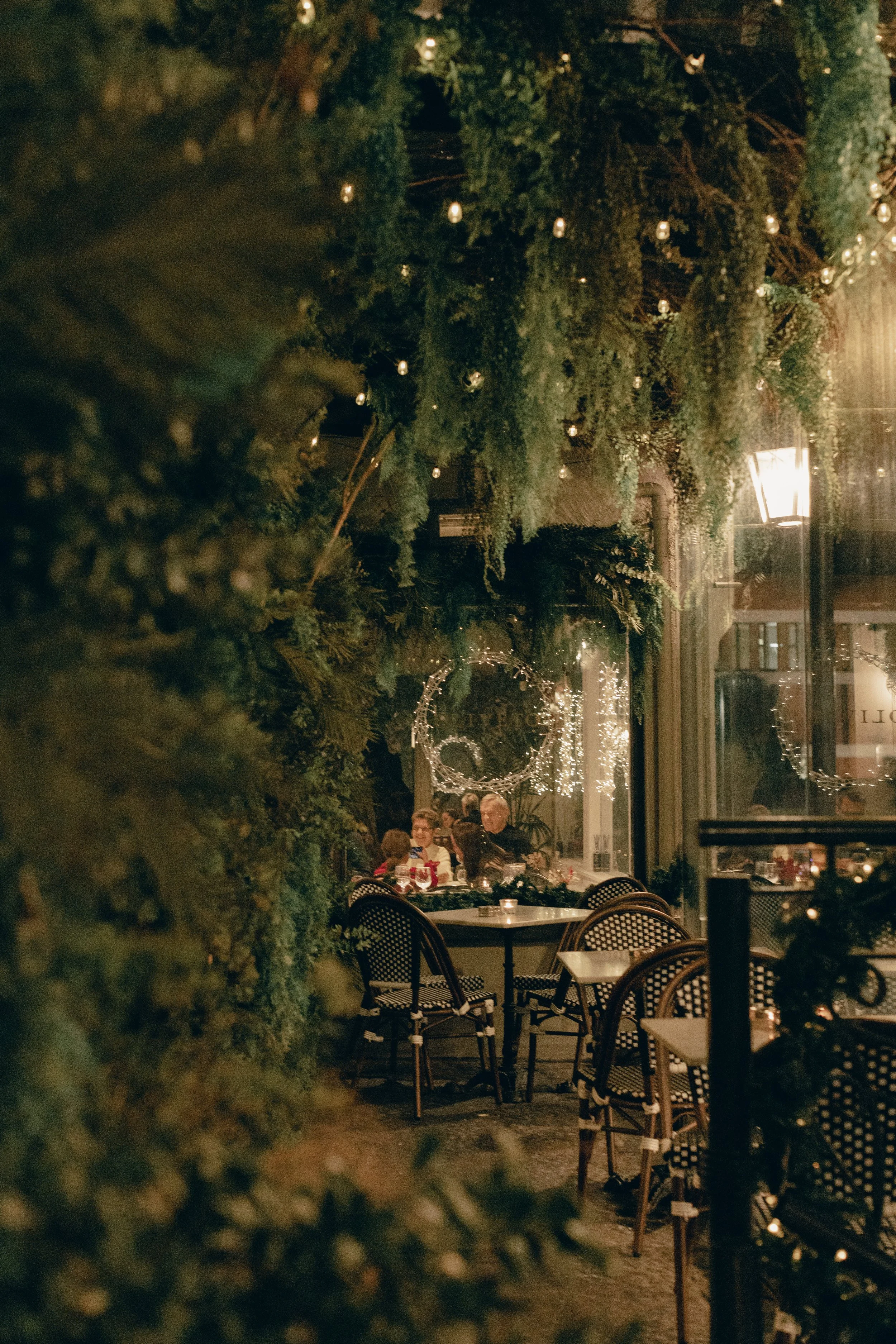 A cozy indoor restaurant with large glass windows showing an outdoor scene decorated with string lights and greenery. Inside, there are tables with guests dining, lit by candles, and an elegant ambiance.