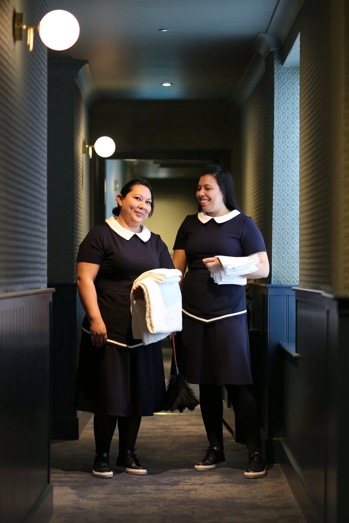 Two housekeepers in navy uniforms with white collars and trim, standing in a hotel corridor, smiling and holding white towels and linens.