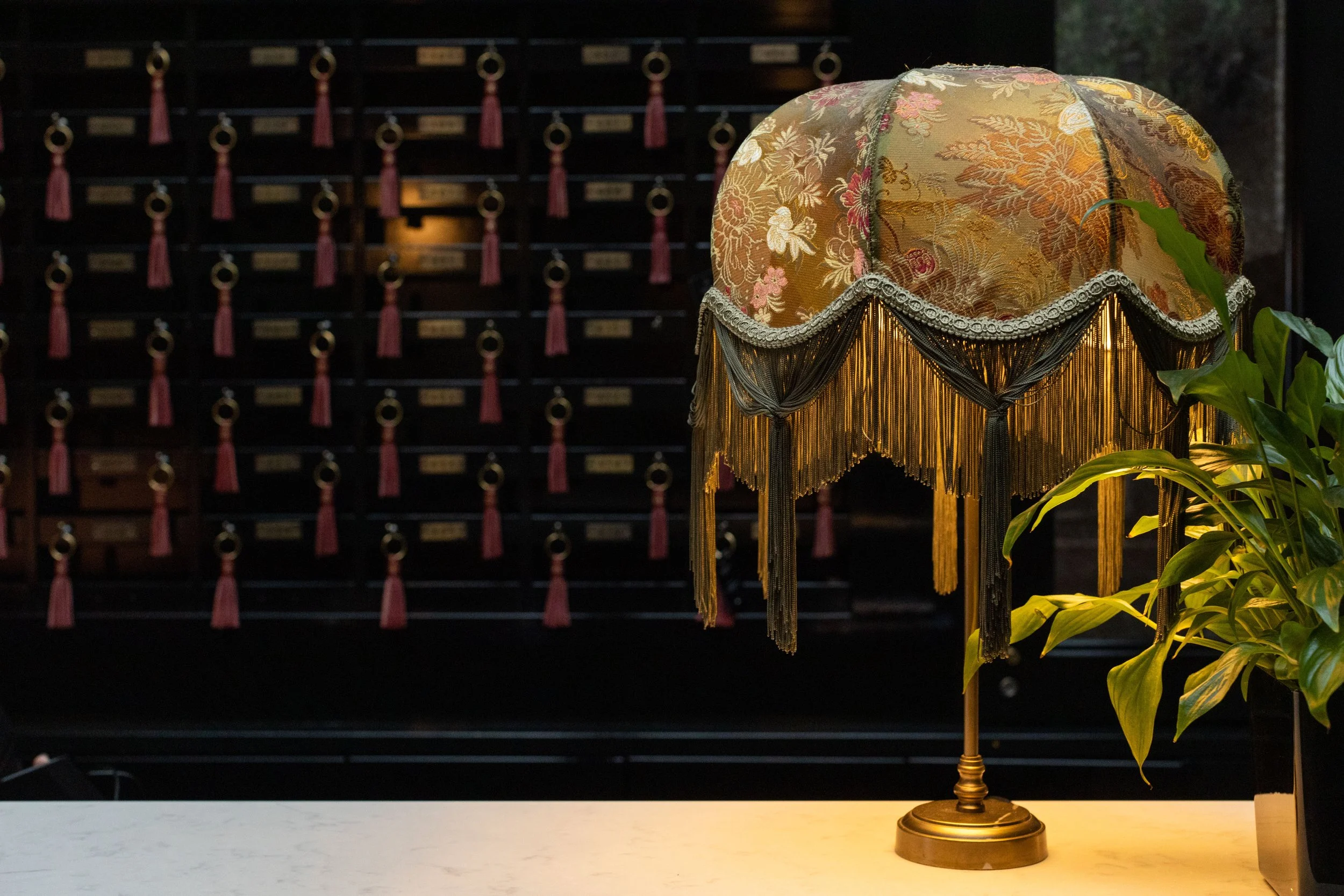 A decorative table lamp with a floral fabric lampshade and beaded fringe, standing on a white surface with a plant nearby. In the background, there is a wooden key holder with small brass plaques and red tassels hanging from hooks.