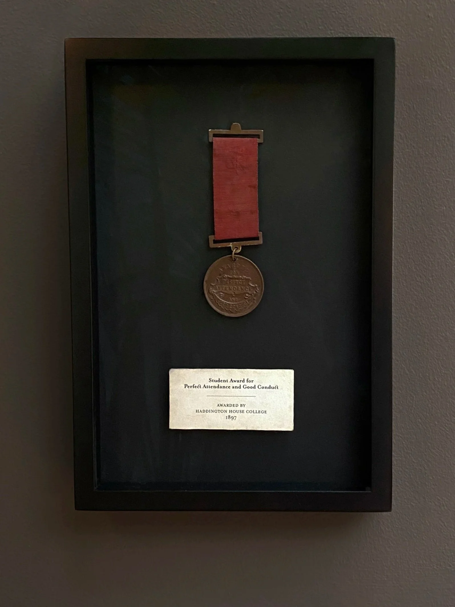Medal with red ribbon and inscription reading 'Perfect Attendance' awarded by Haddington House College in 1897, displayed in black case.