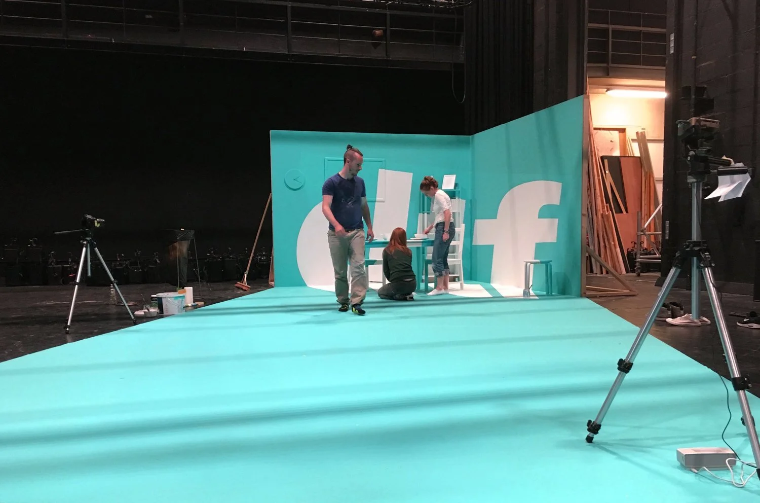A behind-the-scenes view of a photo shoot for Facebook, featuring a blue backdrop with Facebook logos, three people preparing the scene, and photography equipment around.