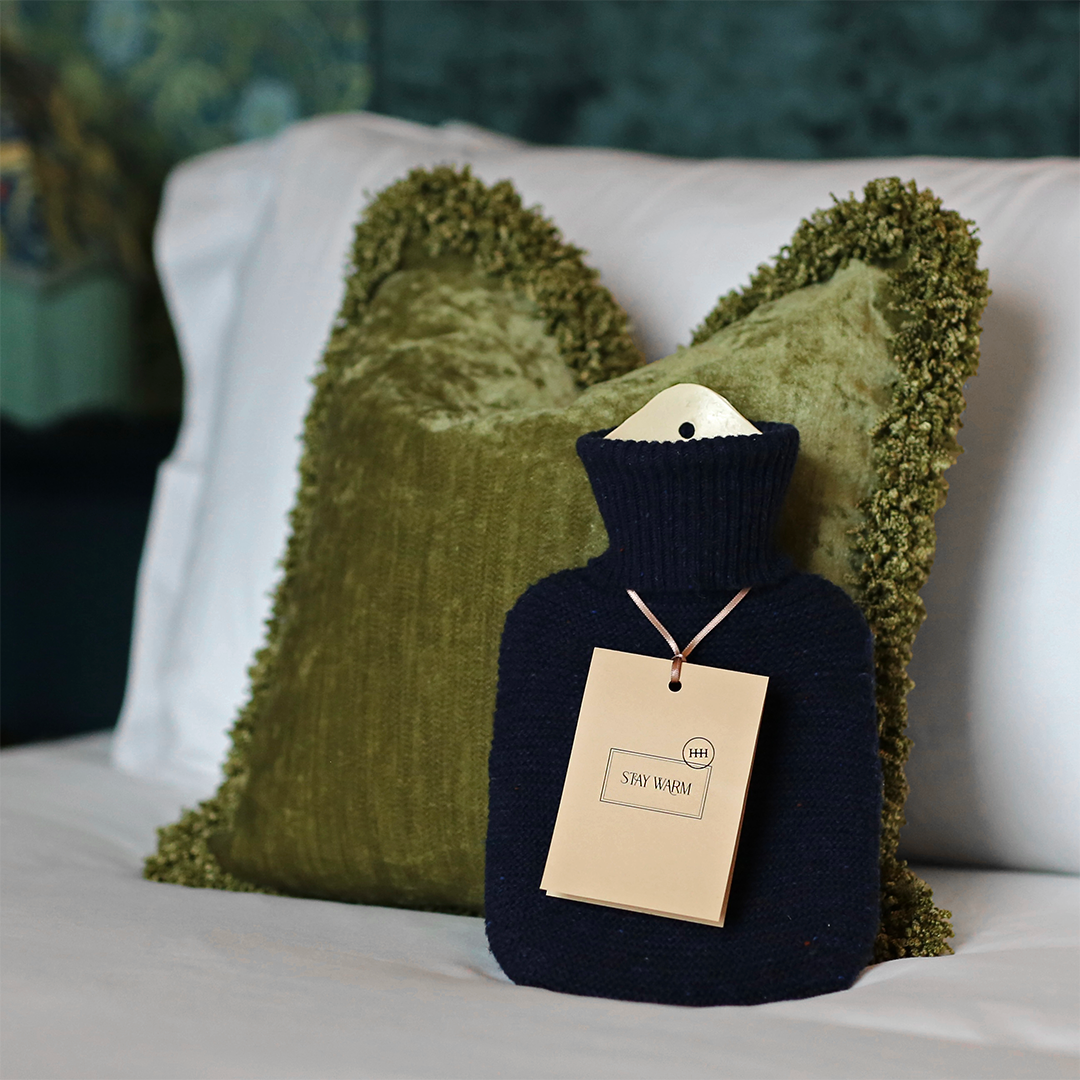 A soothing hot water bottle with a message tag that says "Stay Warm" placed on a bed with two green velvet pillows in the background.