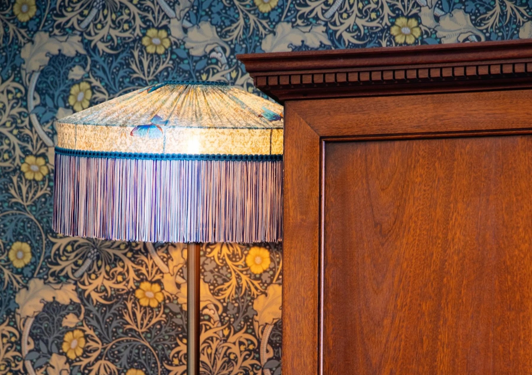 A close-up of a wooden wardrobe beside a decorative standing lamp with a colourful lampshade, against a background of ornate floral wallpaper.