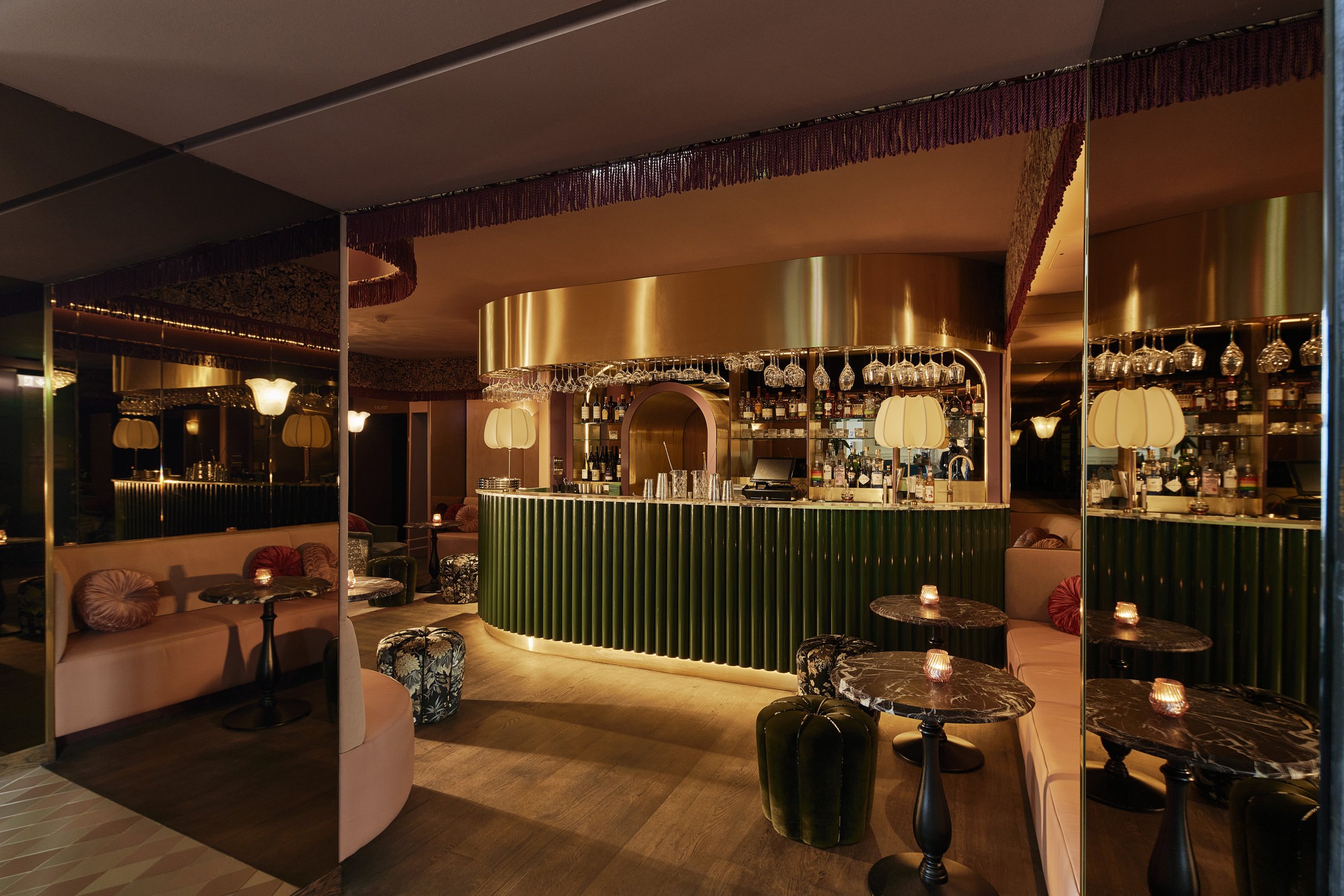 Interior of a stylish bar with a curved green bar counter, glassware hanging above, warm lighting, and plush seating with patterned cushions.