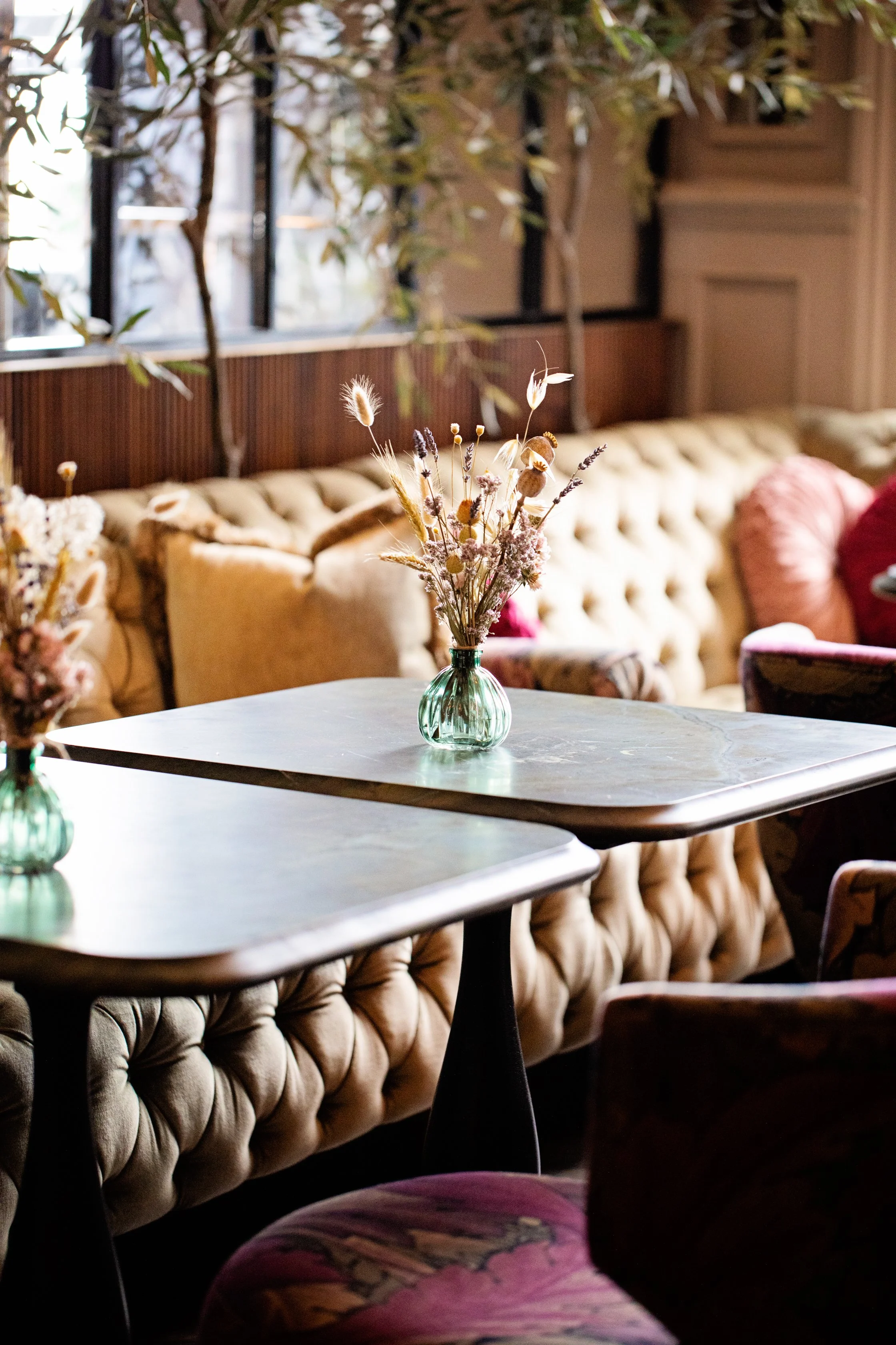 A cozy seating area with tufted beige velvet couches, a round table featuring a glass vase with dried wildflowers, and large windows with black framing letting in natural light, in a warmly decorated interior.