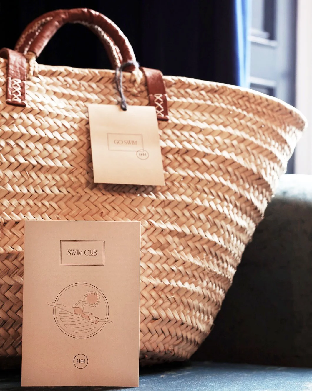 Close-up of a woven straw beach bag with leather handles, with tags and a paper card that reads 'Swim Club' with a seal and a line drawing of a swimmer.