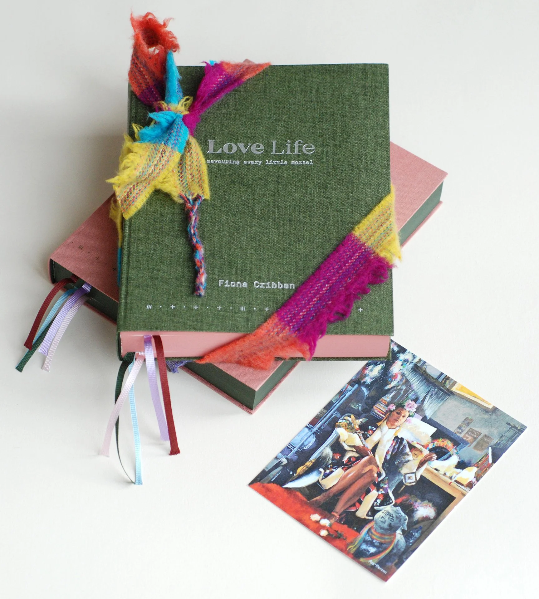 A green felt-covered book titled 'Love Life' by Flona Cribben, decorated with colorful knitting and ribbons, resting on top of a pink book. A photo of a colorful city scene is partially visible beneath them.