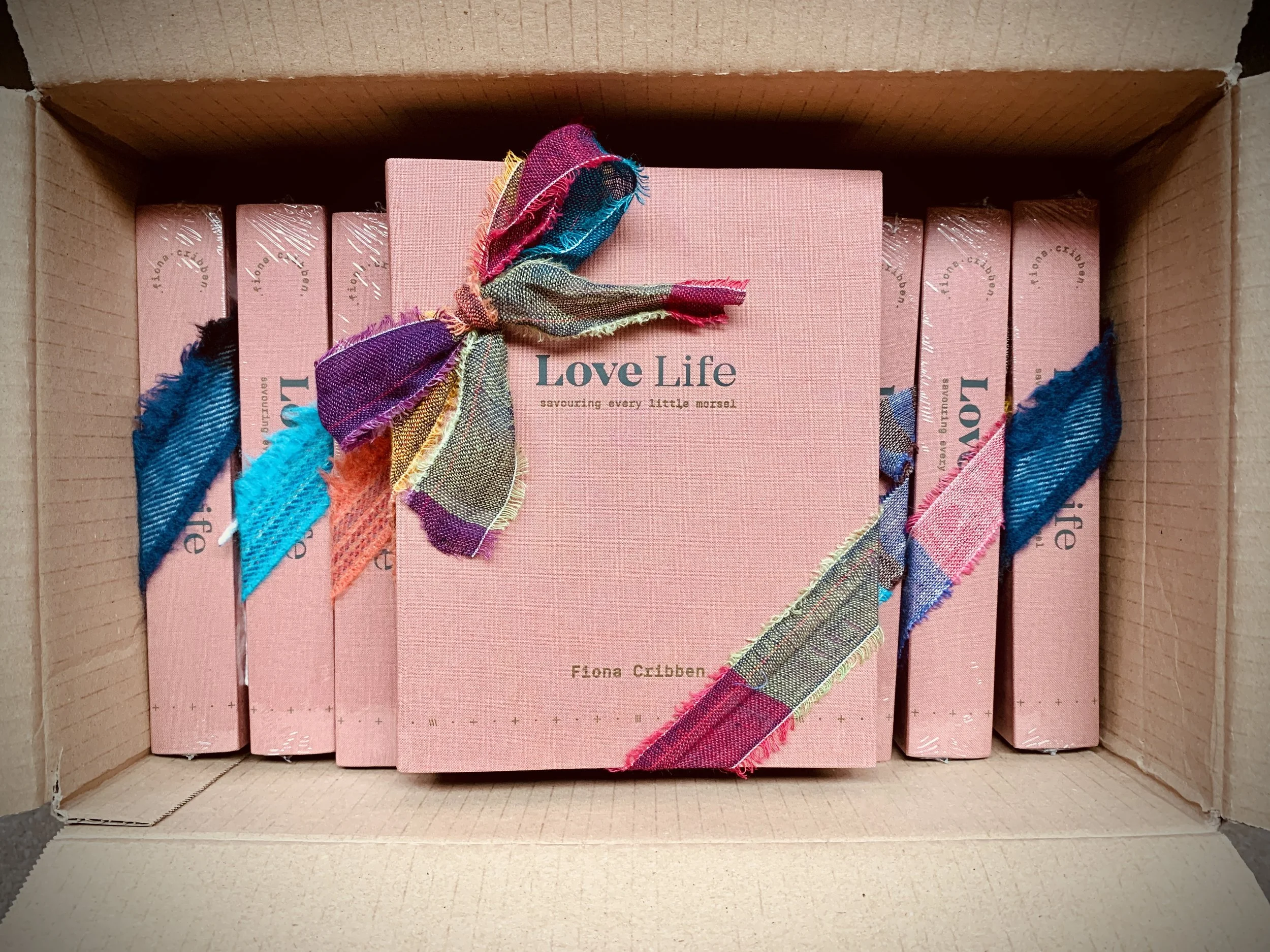 A cardboard box containing a pink book titled 'Love Life' by Fiona Cribben, decorated with multicolored fabric ribbons.