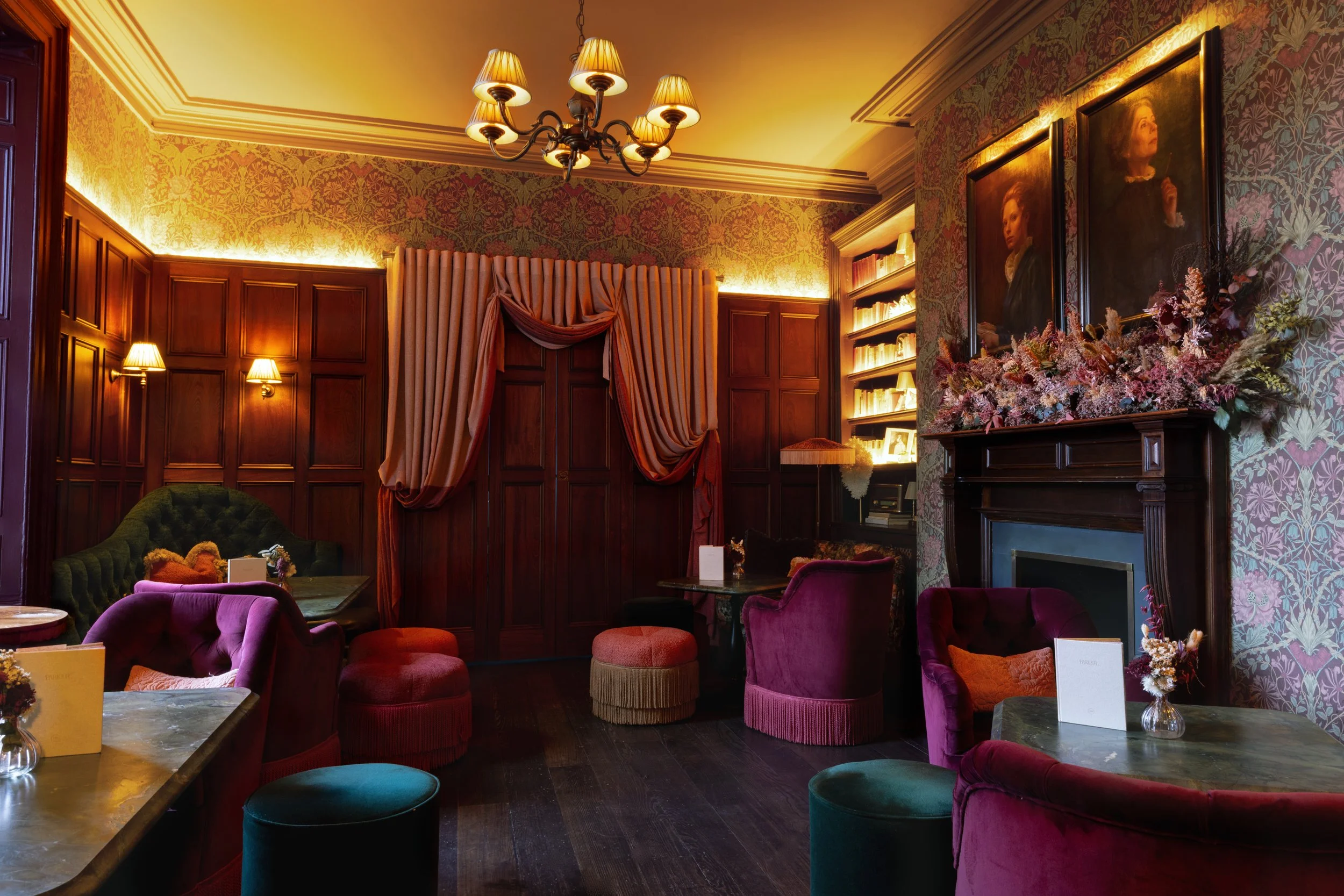 Elegant lounge with purple, green, and pink velvet chairs, wooden panel walls, floral wallpaper, paintings, a fireplace decorated with flowers, and warm lighting from wall sconces and a chandelier.