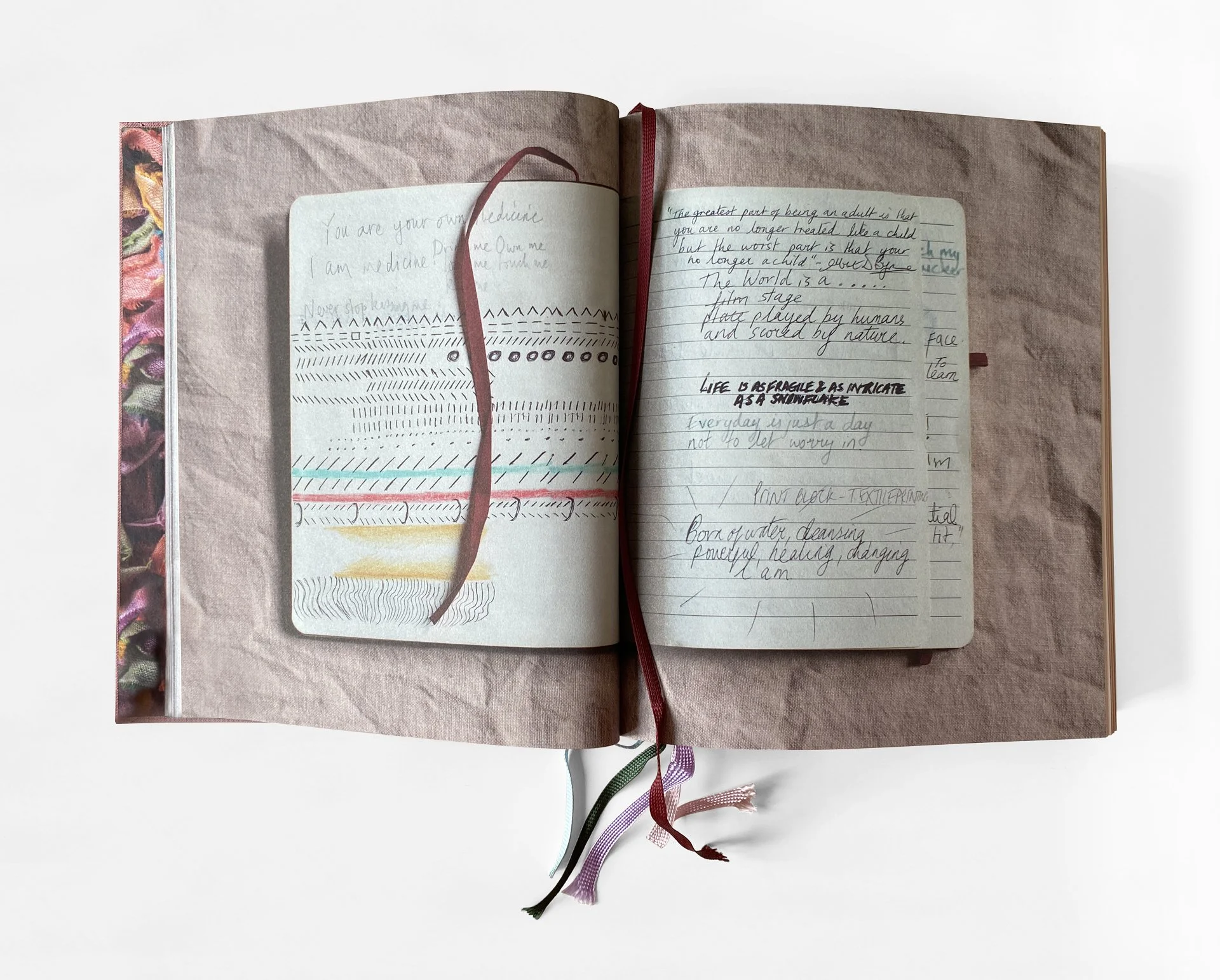 Open notebook with handwritten notes and a colorful fabric cover visible on the left. The pages contain cursive writing and decorative lines, with multiple ribbons hanging from the binding.