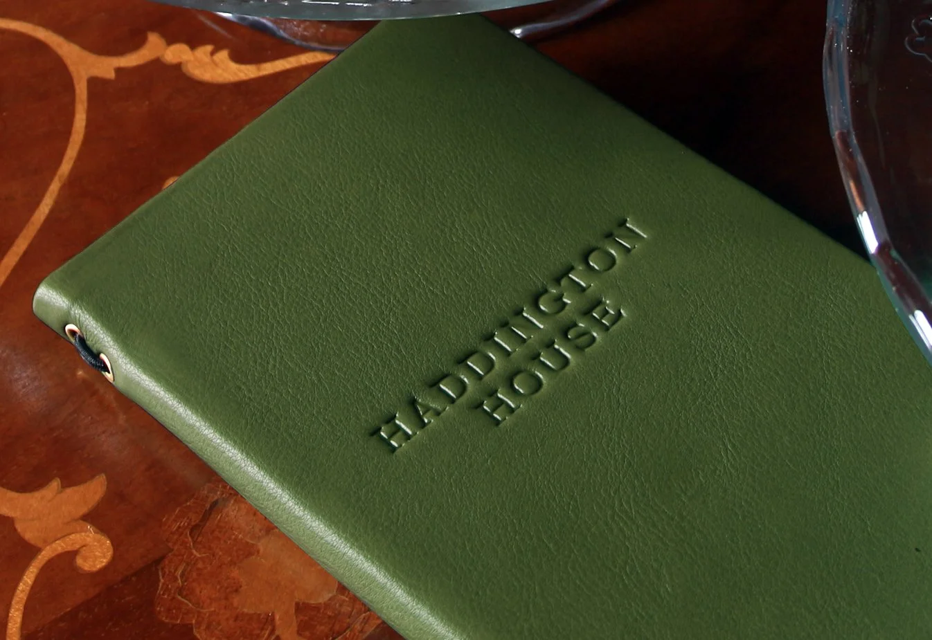 Close-up of a green leather-bound menu with embossed text reading 'HADDINGTON HOUSE' on the cover, placed on a wooden table.