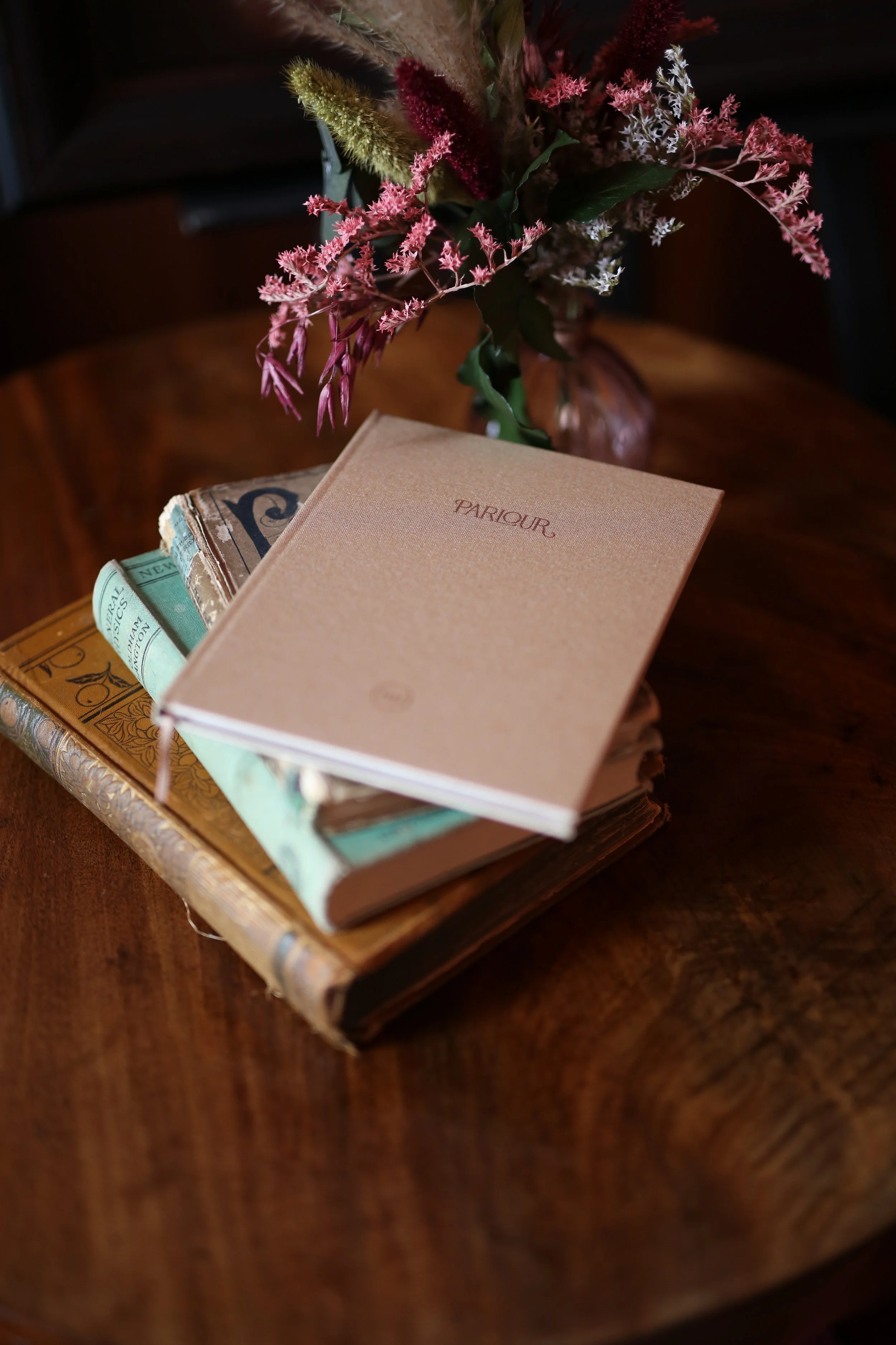 A small bouquet of dried flowers and grasses in a vase behind several stacked books on a wooden surface.