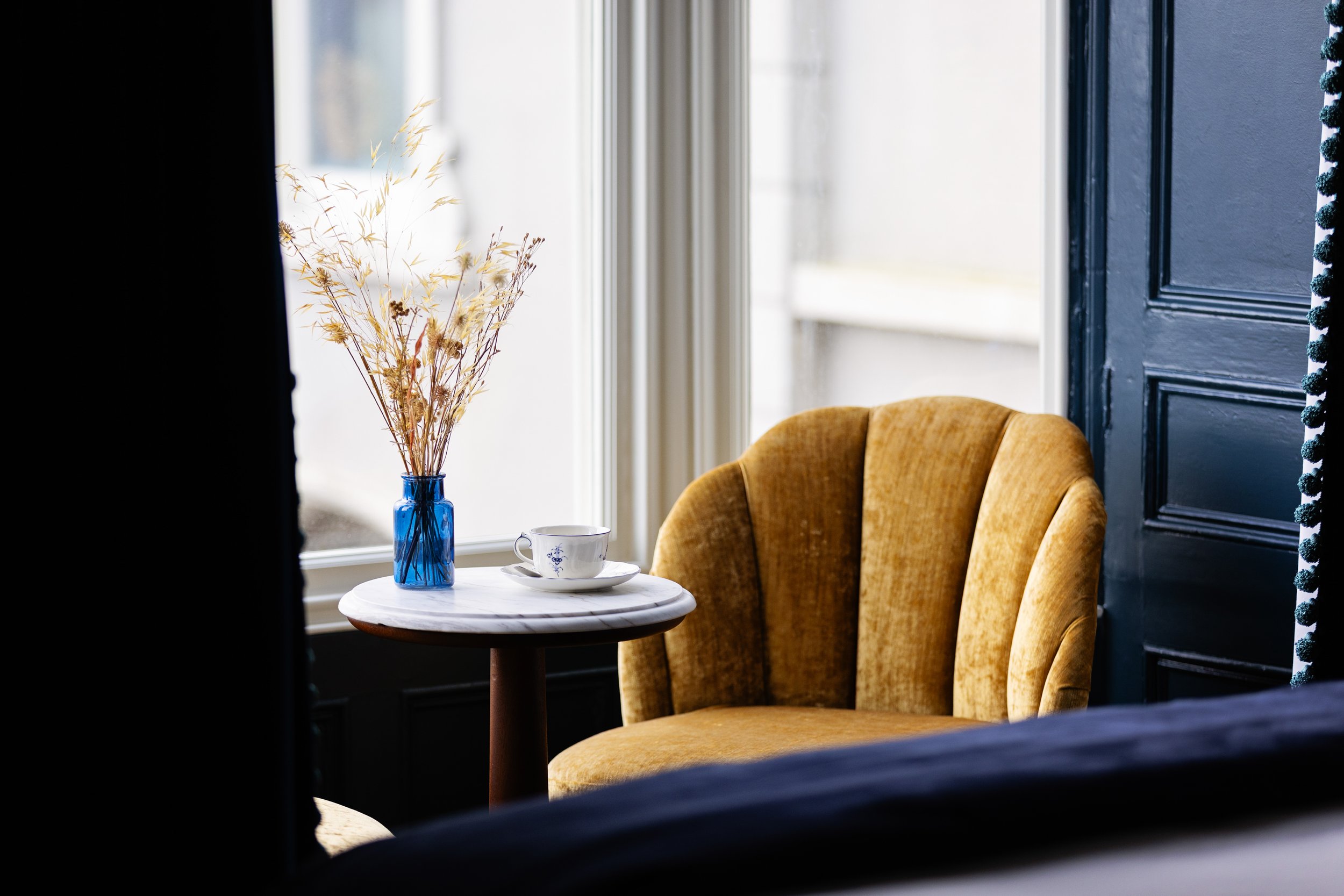 A cozy corner with a round marble table holding a blue vase with dried flowers, a china teacup, and an armchair with yellow upholstery near a window and a dark door.
