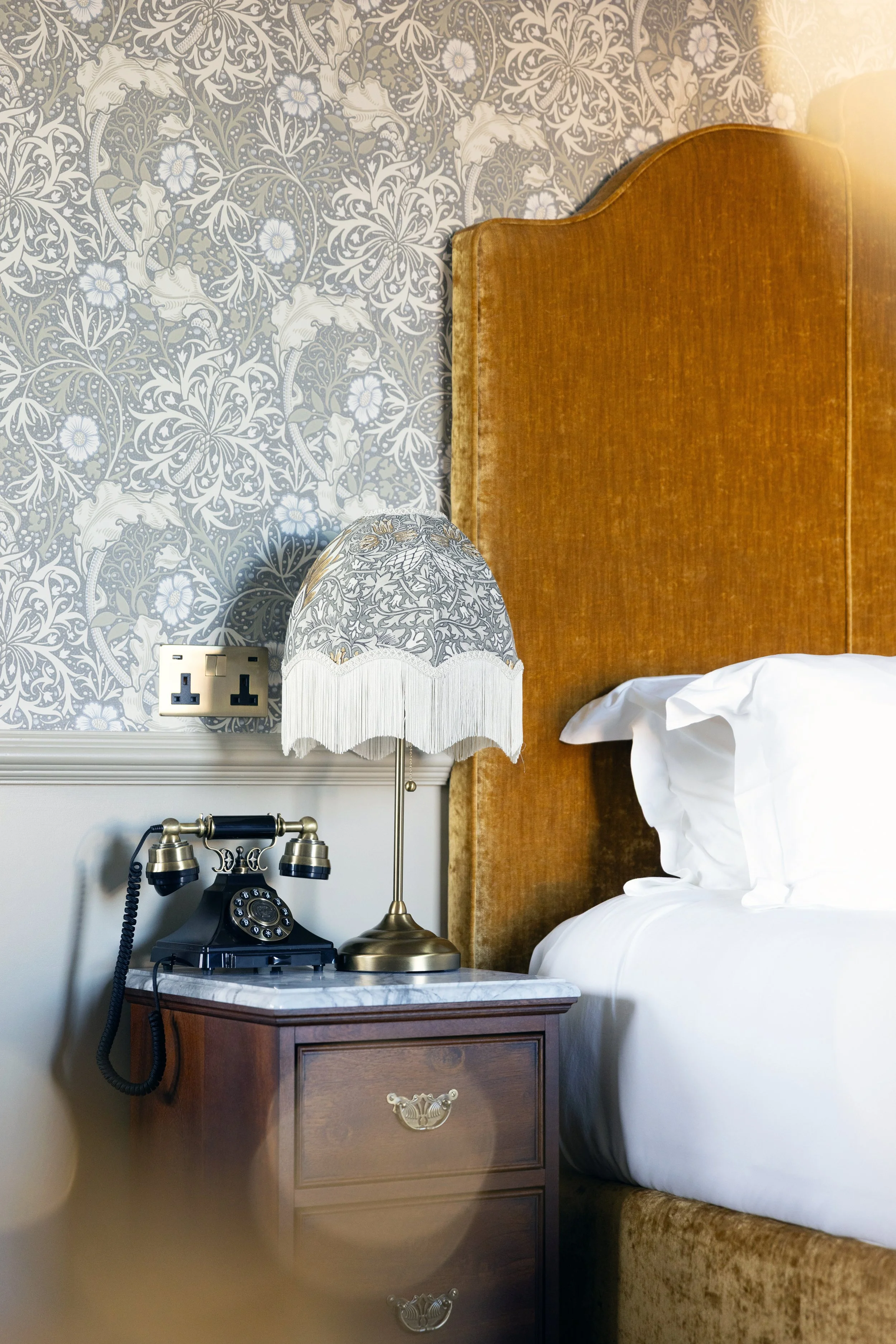A bedside table with a vintage black rotary phone, a brass bedside lamp, and a lampshade with a floral and animal pattern. The headboard is brown wood, and the wall features patterned wallpaper with floral and lion motifs.