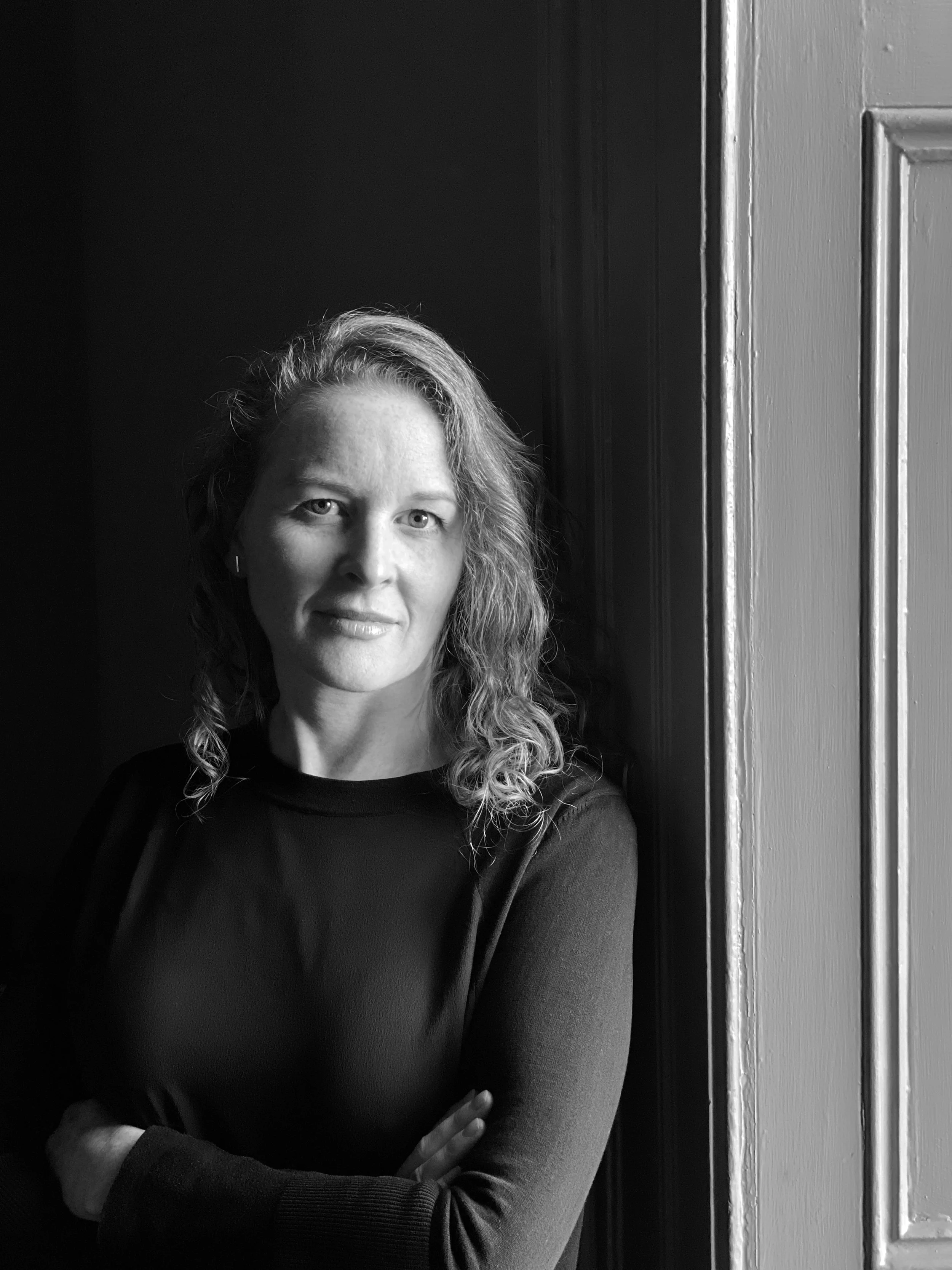 Black and white portrait of a woman with wavy hair, standing near a window with her arms crossed.