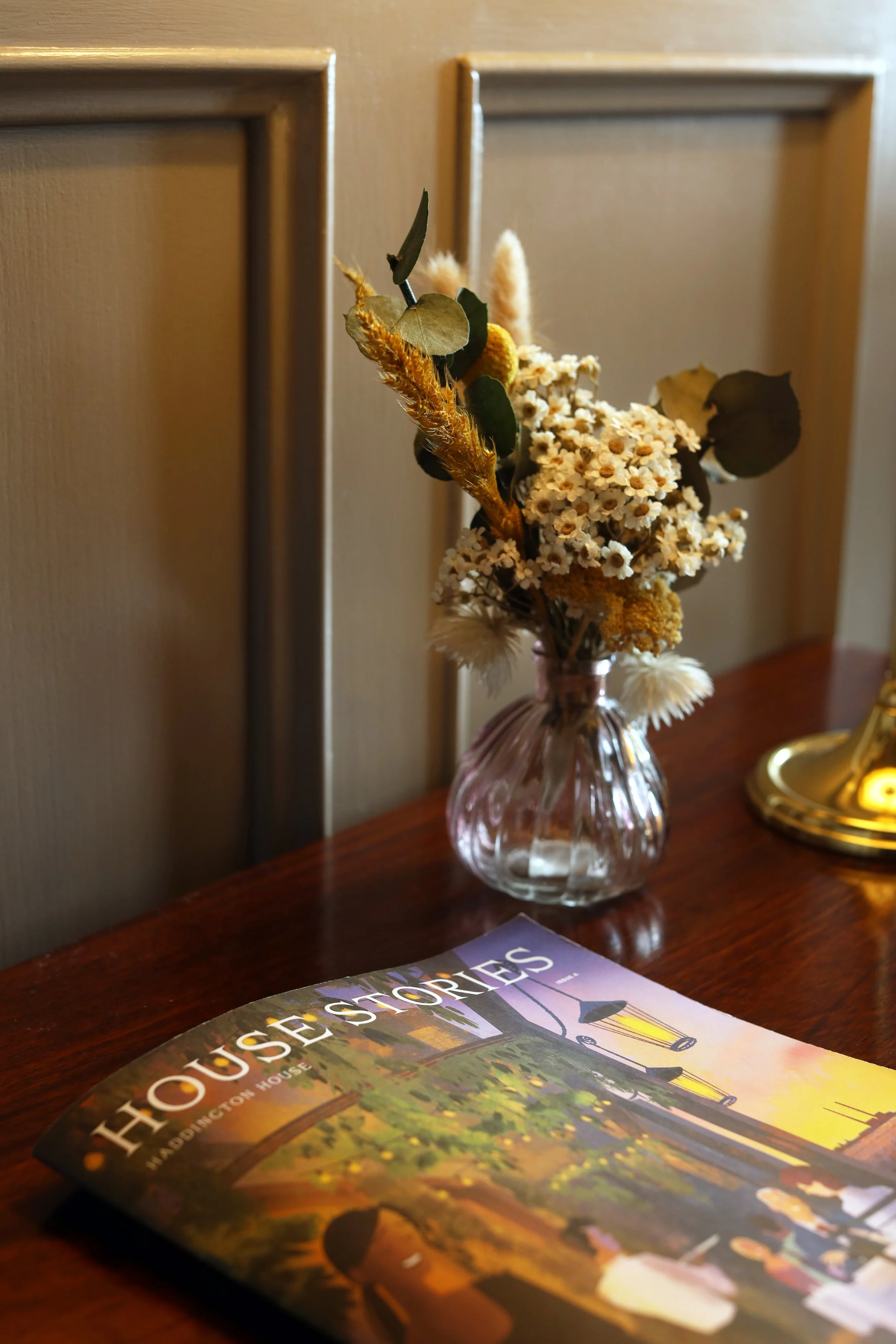 A bouquet of dried flowers in a pink glass vase on a wooden table, with a travel magazine in the foreground and a wooden paneled wall in the background.