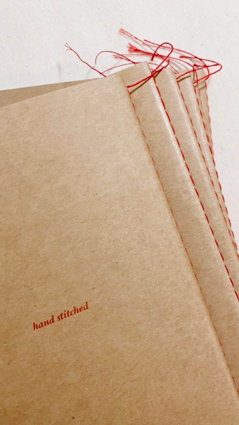 A close-up of a stack of beige paper bags with red stitching and red thread ties, with the words 'hand stitched' printed on one of the bags.