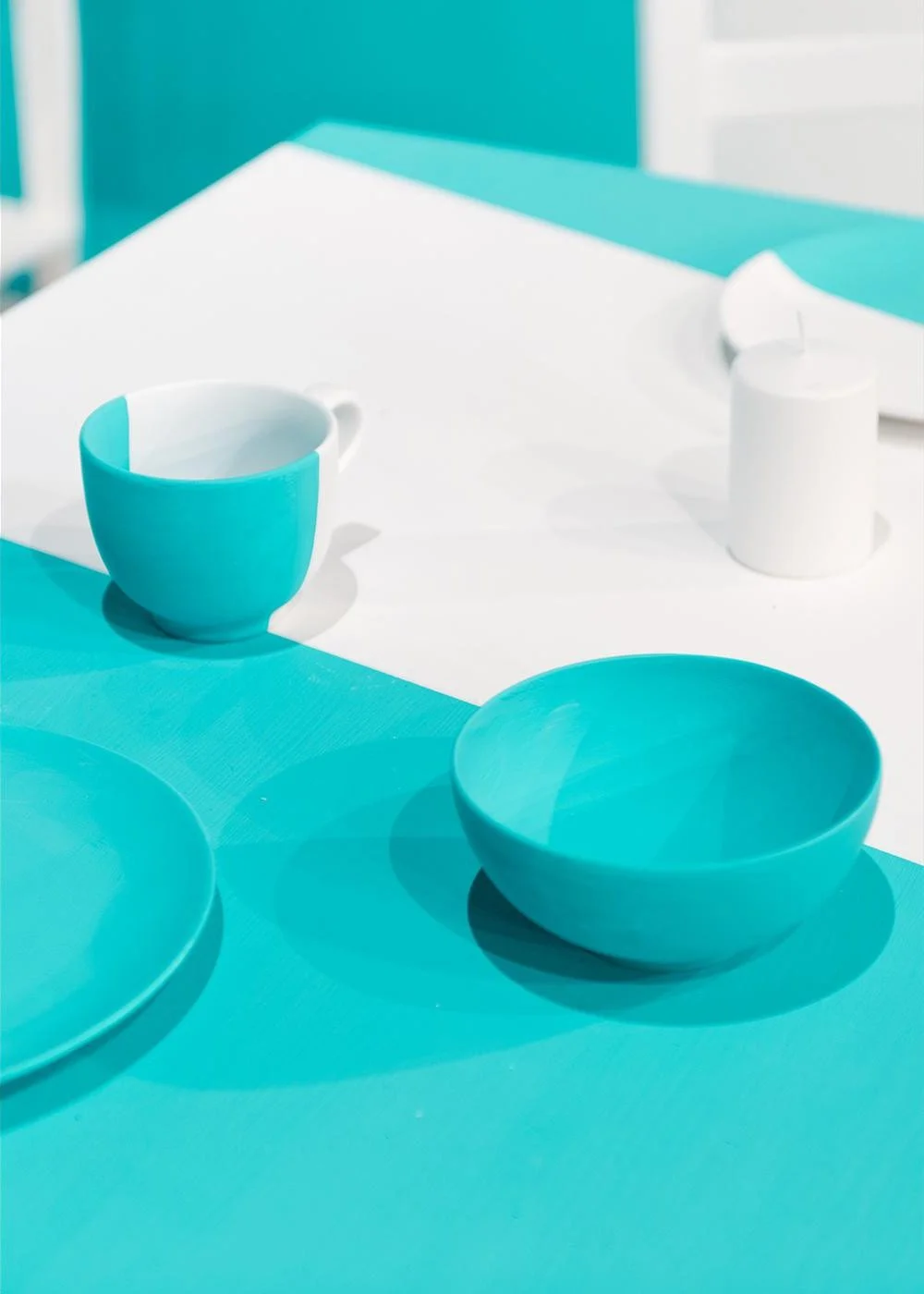 Turquoise and white tableware, including a cup, bowls, and a mug, arranged on a white and turquoise table with shadows.