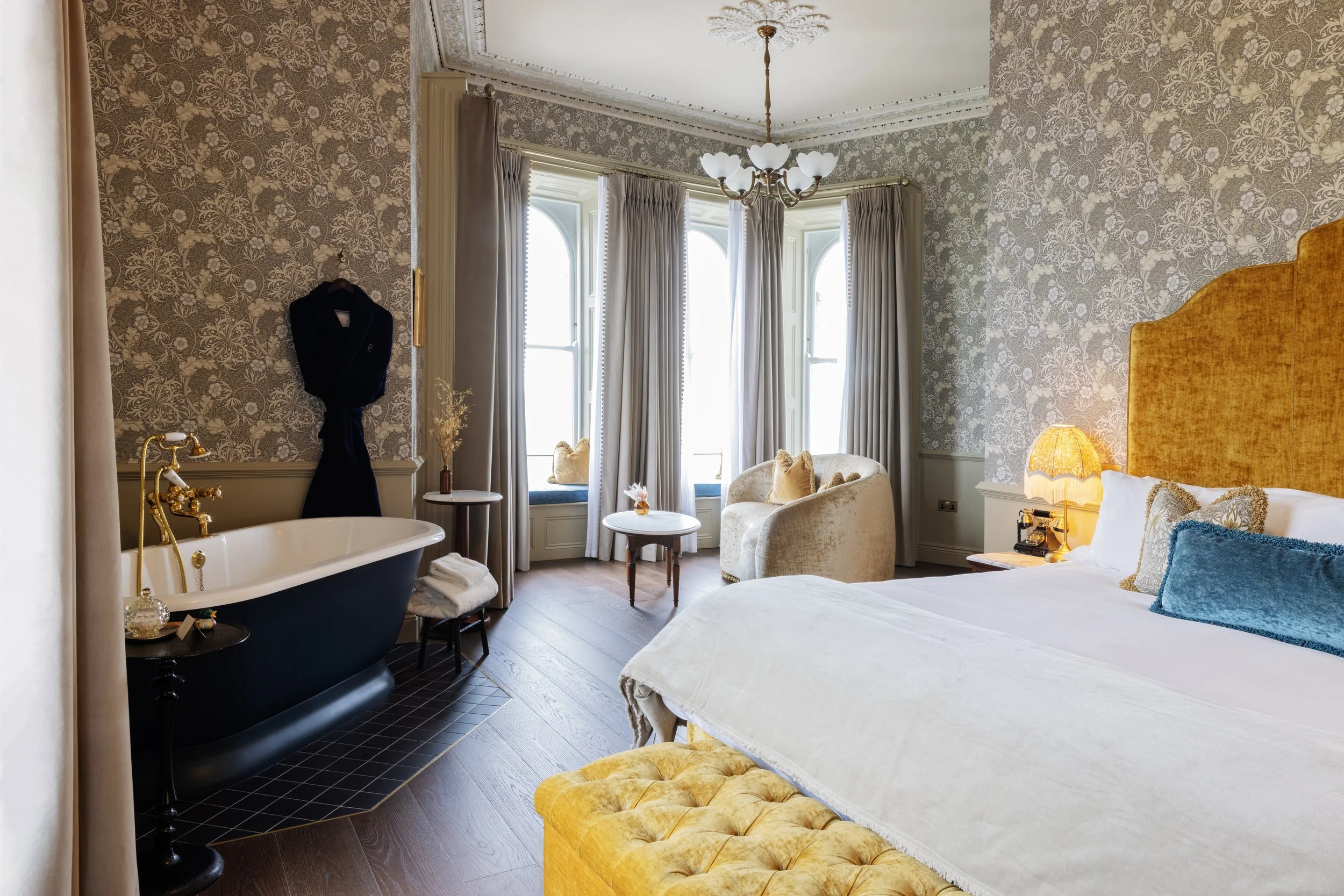 A hotel room with two beds, ornate wallpaper, a chandelier, a vintage bathtub, a sitting area with chairs and a table, large windows with curtains, and warm lighting.