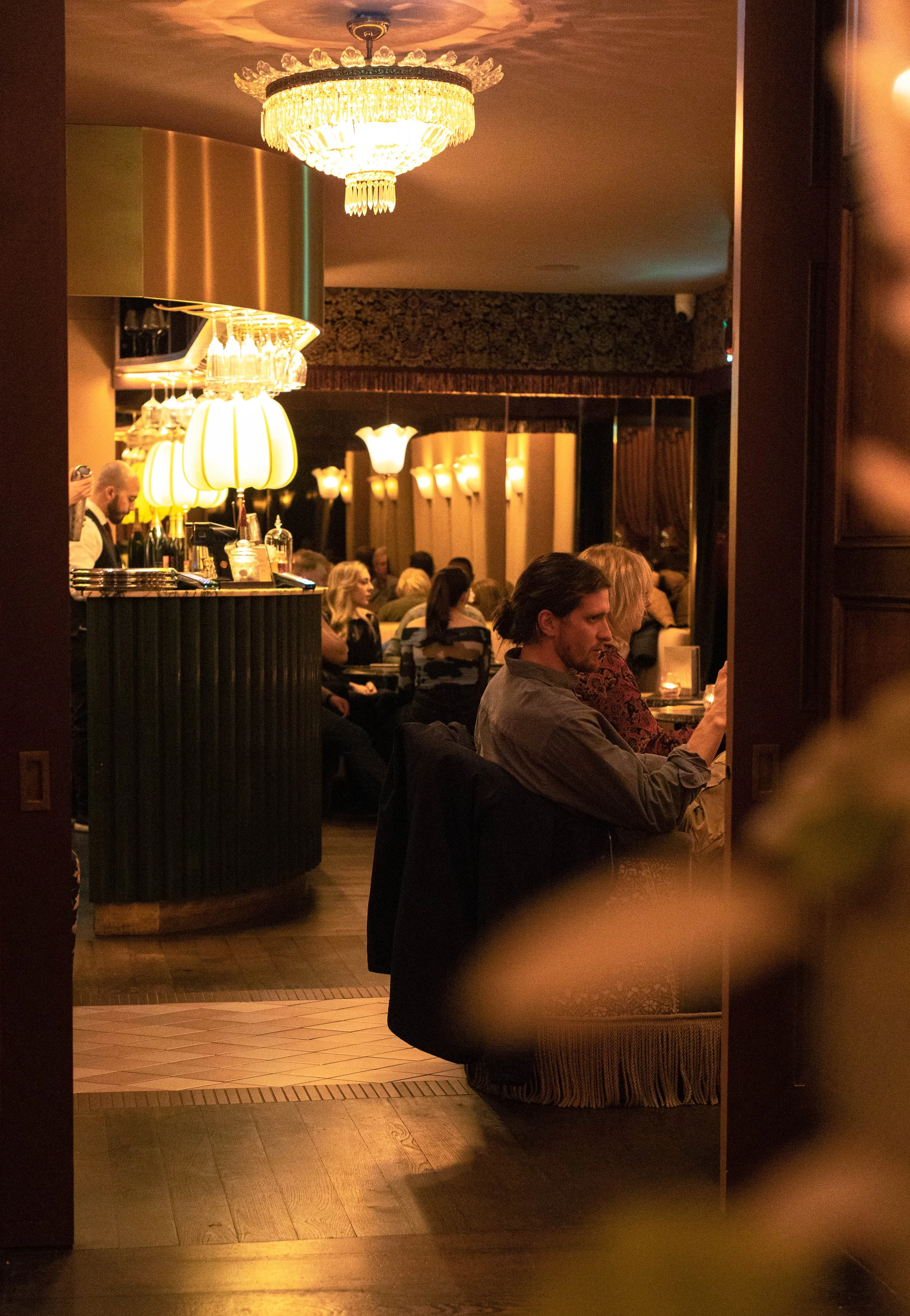 A view of a cozy restaurant or lounge with a large, ornate chandelier hanging from the ceiling. The room is filled with warm lighting, and there are several people seated at tables, some engaged with their phones or conversing. The scene appears to be in the evening, creating an intimate atmosphere.