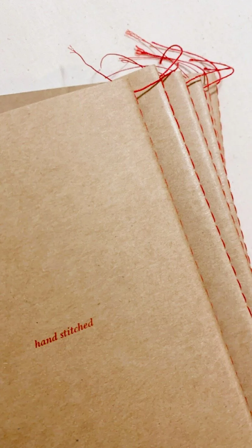 A stack of brown paper pages with red sewing stitches at the edges and red thread frays, with the words "hand stitched" printed on one page.