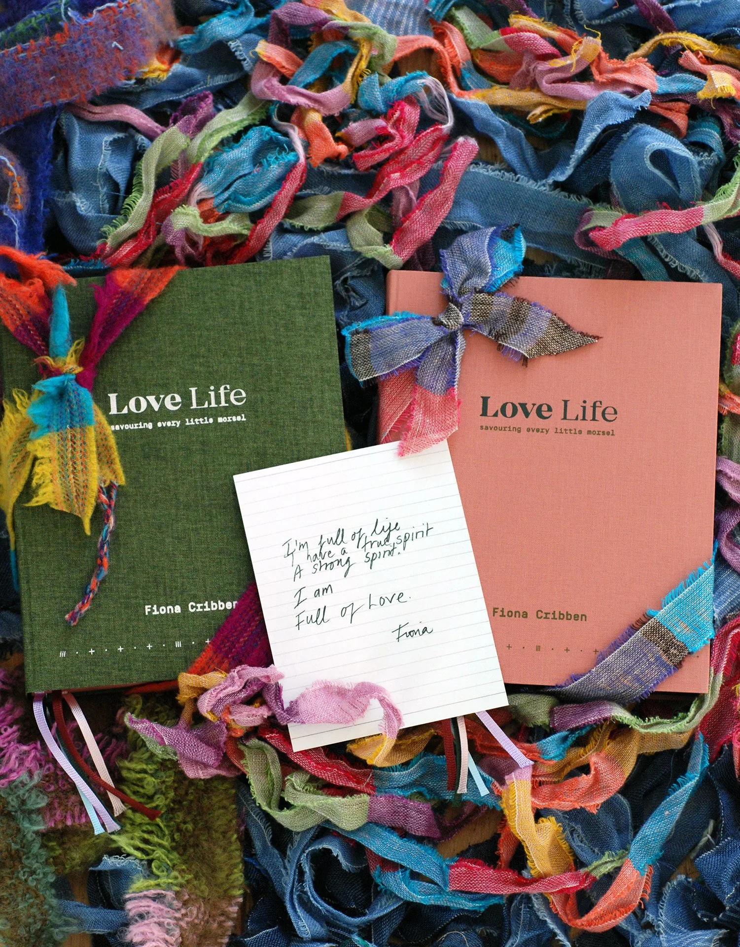 Close-up of two books titled "Love Life" by Fiona Cribben, surrounded by colorful, intertwined fabric strips and a handwritten note read by someone named Fiona.