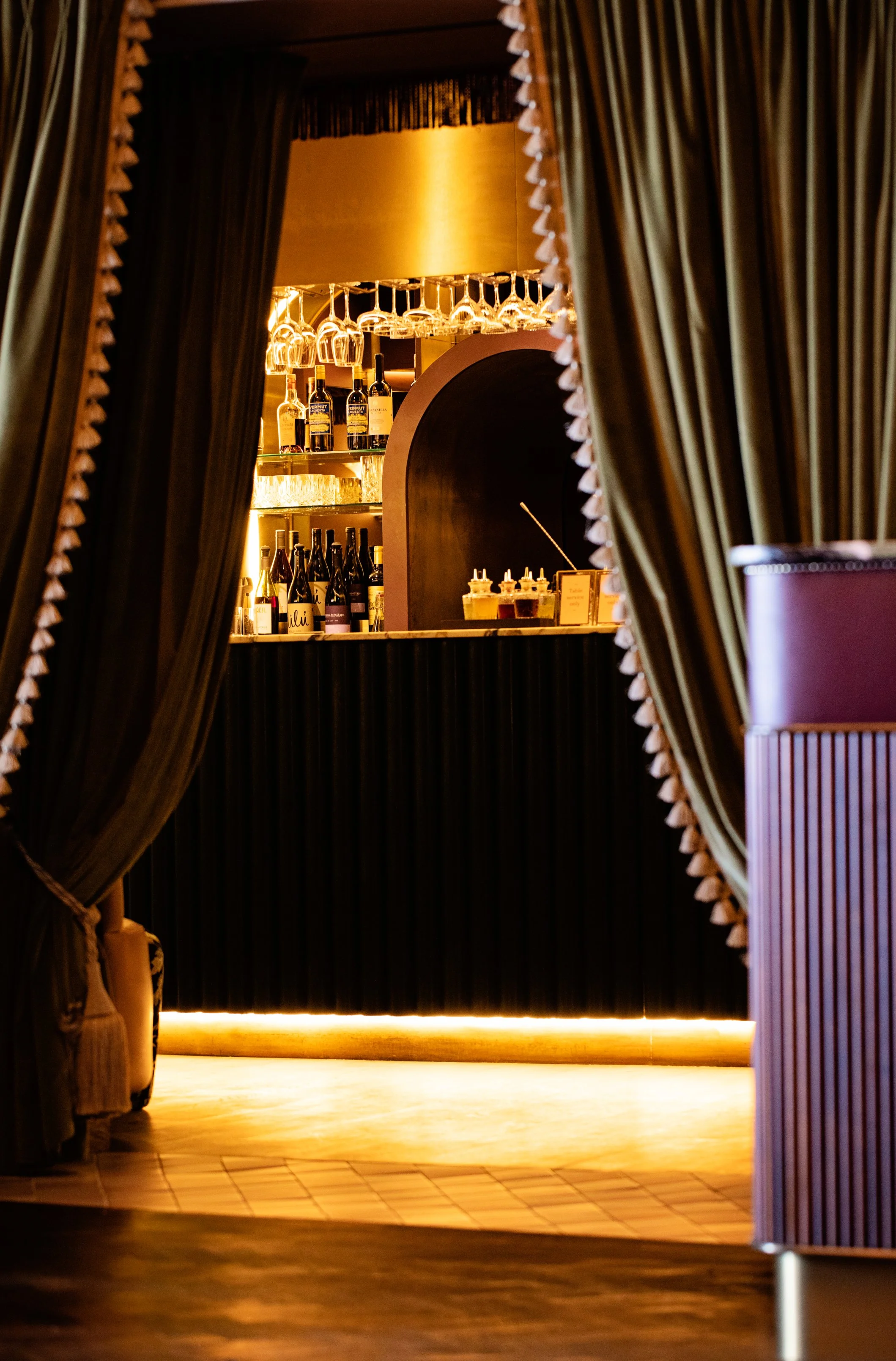 View through curtains revealing a bar area with wine bottles and hanging glasses, illuminated by warm ambient lighting.