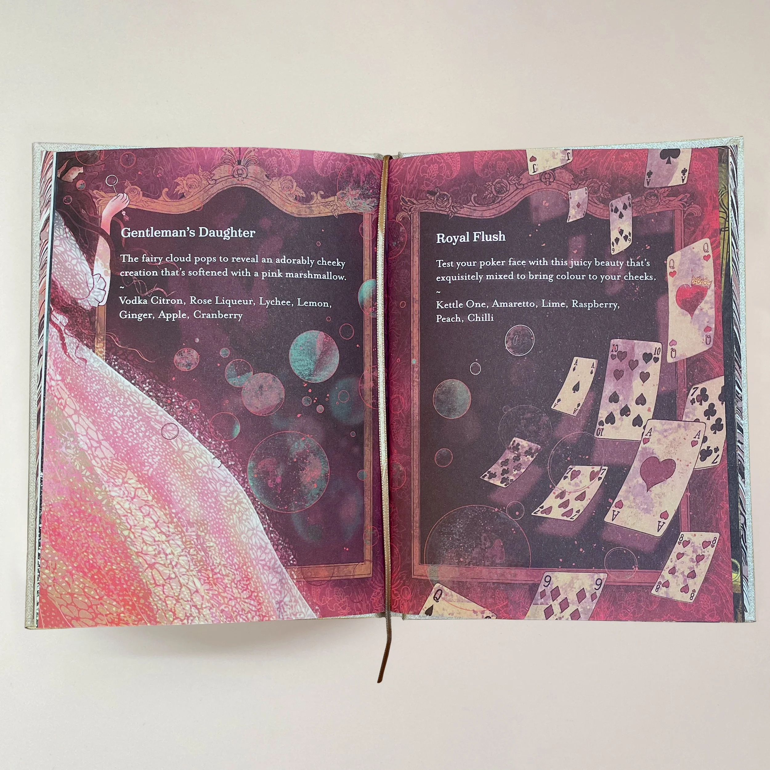 Open magazine showing two pages with text and illustrations, decorated with pink and purple artwork, including floating playing cards and bubbles, on a white background.