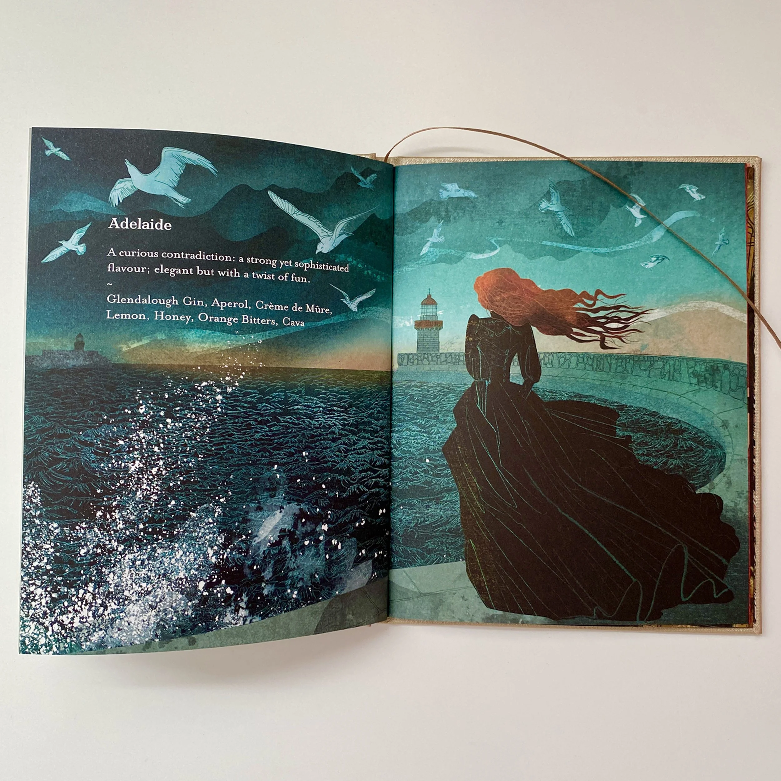 Open book featuring a colorful illustration of a coastal scene with seagulls flying above the sea, a lighthouse, and a rocky shoreline with a person standing on it.