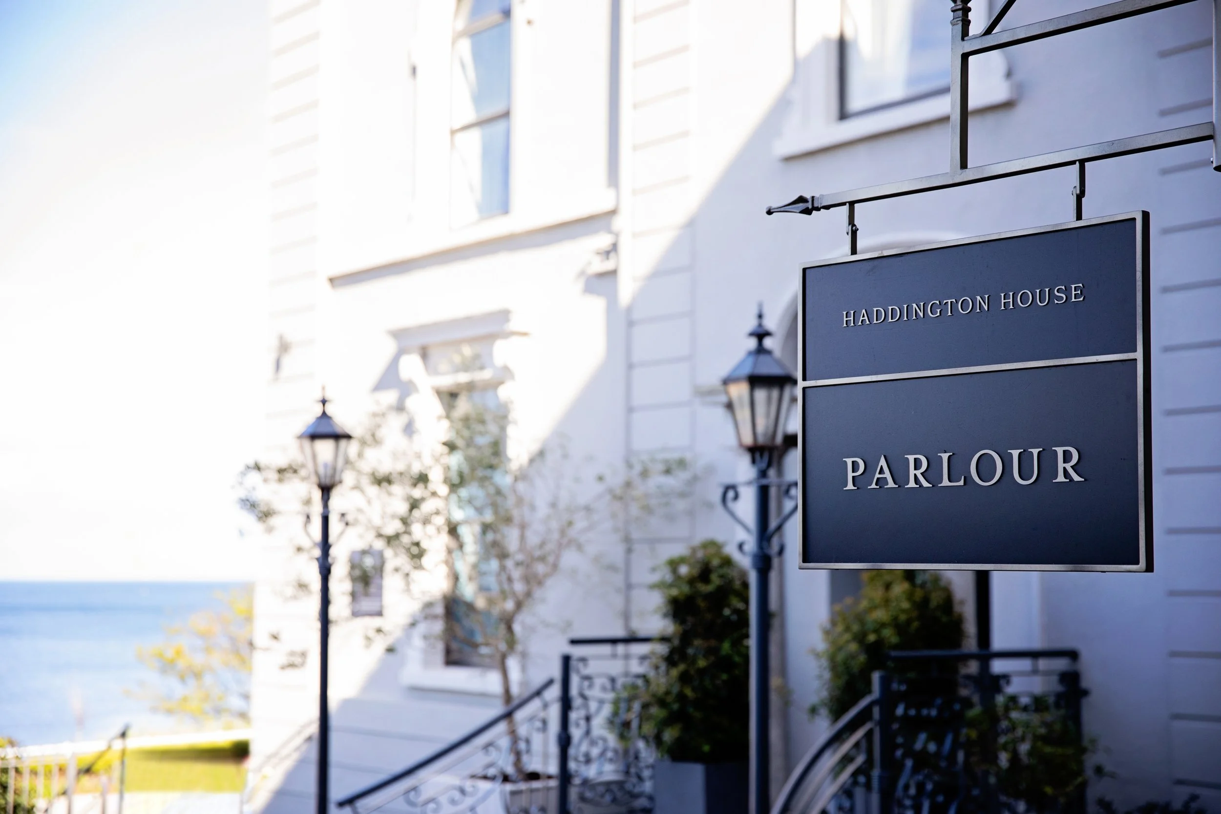 A sign for Haddington House Parlor hangs outside a white building with multiple windows and street lamps, near a waterfront or ocean view.