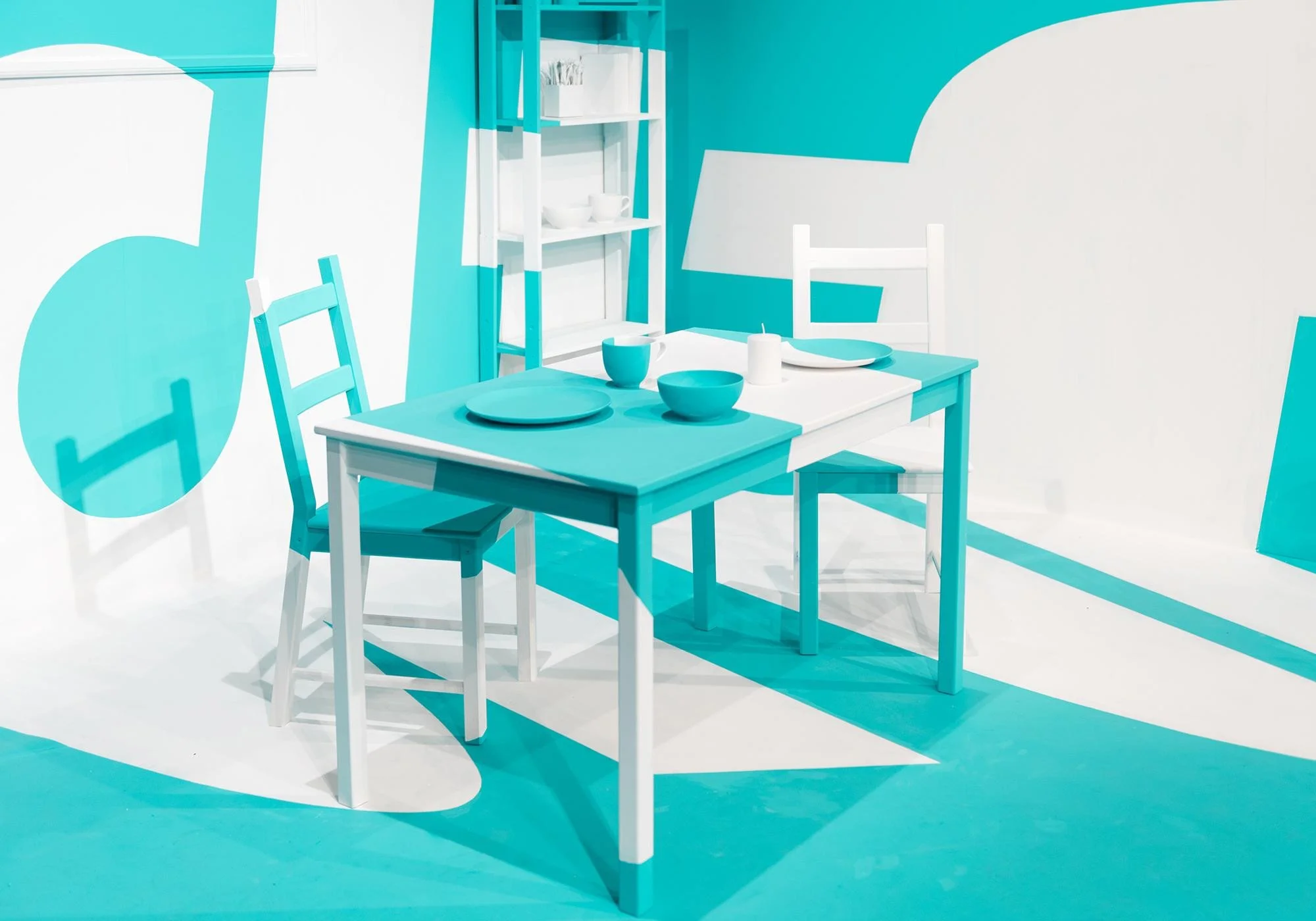 A bright, modern dining area with turquoise and white color scheme. Includes a white table with turquoise placemats, bowls, and cups, surrounded by white chairs. The wall has turquoise and white abstract shapes, and a turquoise and white shelving uni