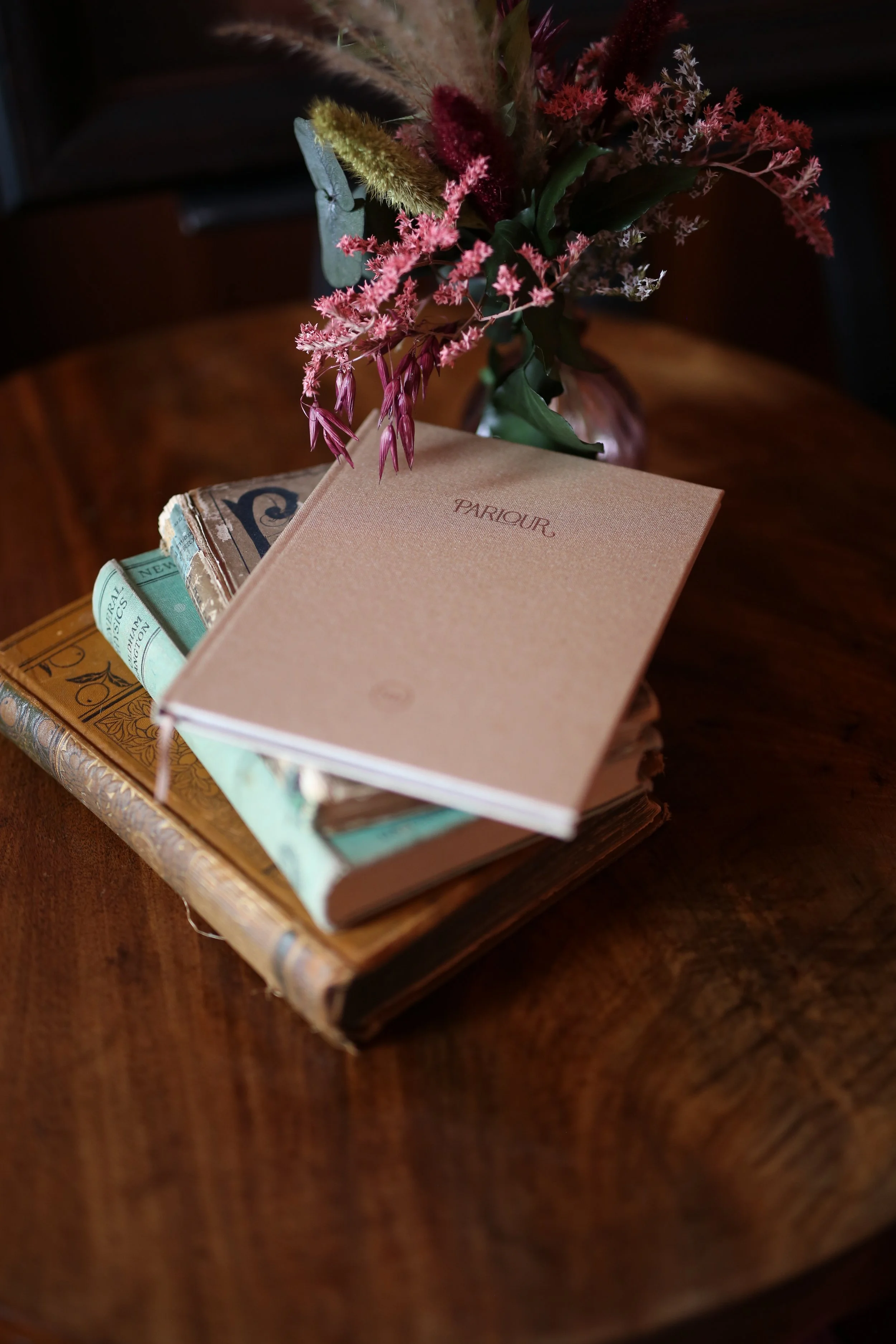 A bouquet of pink, yellow, and white flowers resting on top of a stack of vintage books on a wooden surface.