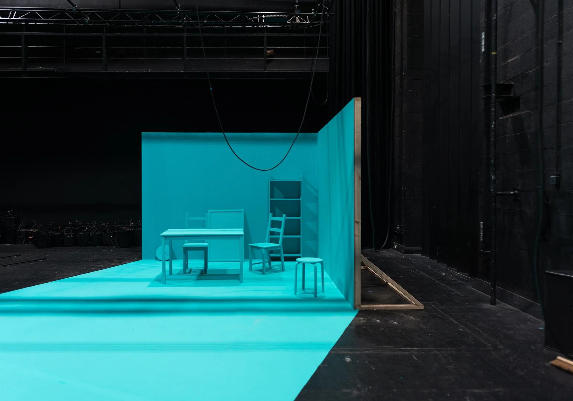 A backstage view of a theater set with a turquoise stage and backdrop. There are four white chairs on the stage, and a small turquoise table. The stage is surrounded by black walls and rigging equipment overhead.