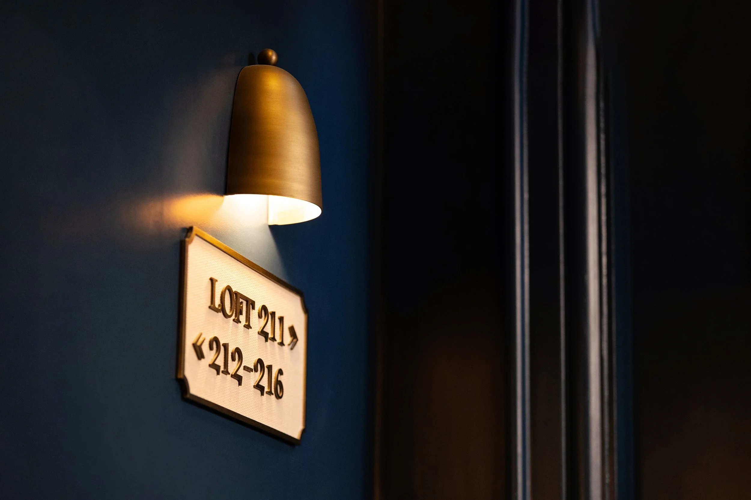 Close-up of a wall-mounted light fixture illuminating a sign with brass text on a beige background, partially visible, showing room numbers at a hotel.