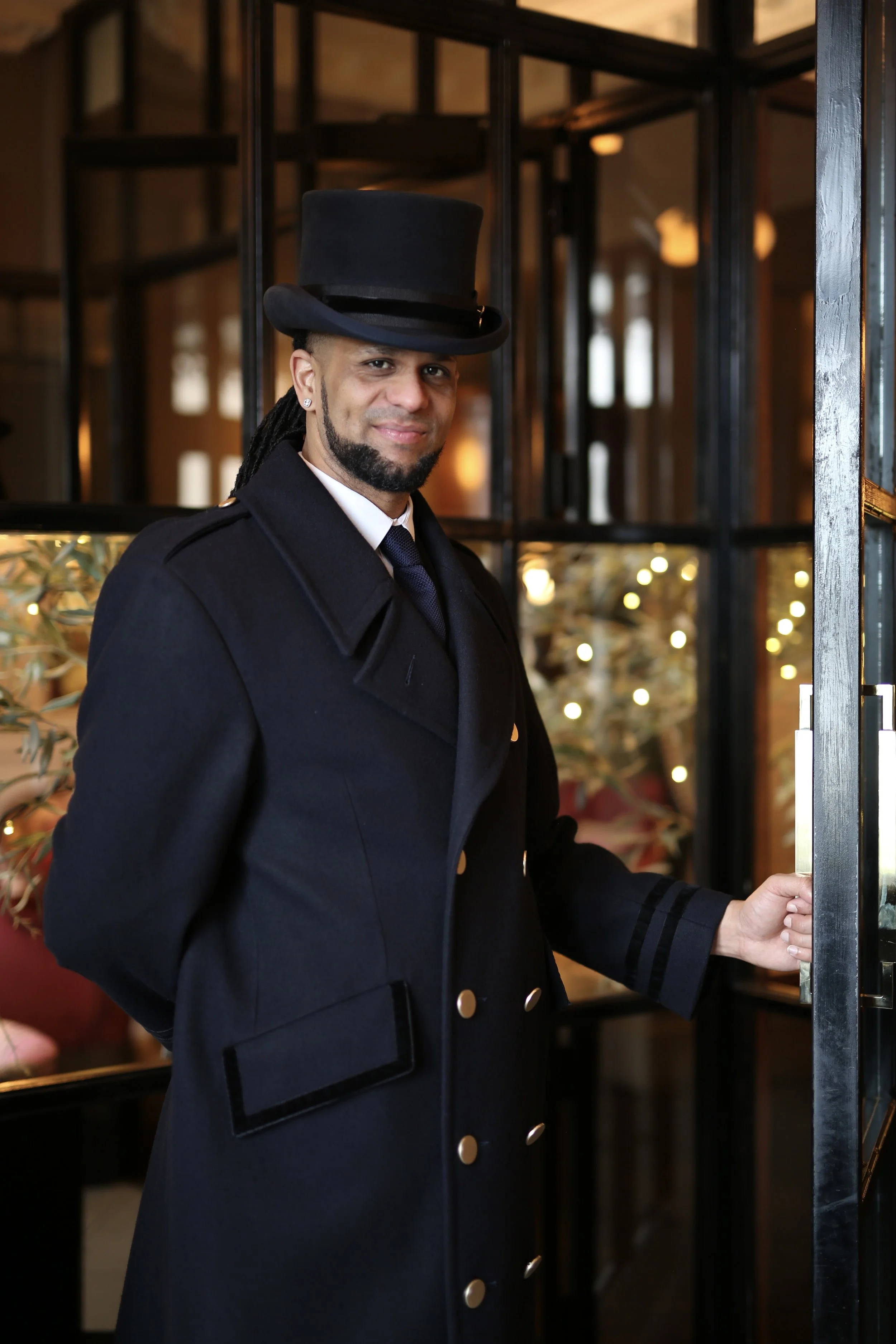 A man dressed in a formal navy blue uniform with a matching gold-button coat and a black top hat standing inside a warmly lit hotel lobby, holding a door handle.
