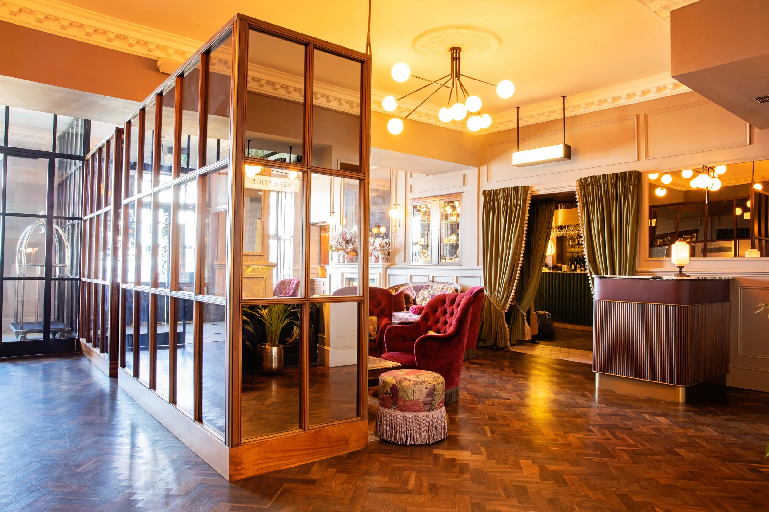 Interior of a cozy lounge or restaurant with wooden flooring, vintage red velvet armchairs, gold curtains, a modern chandelier, and decorative lighting. Glass partitions separate seating areas, creating an inviting atmosphere.