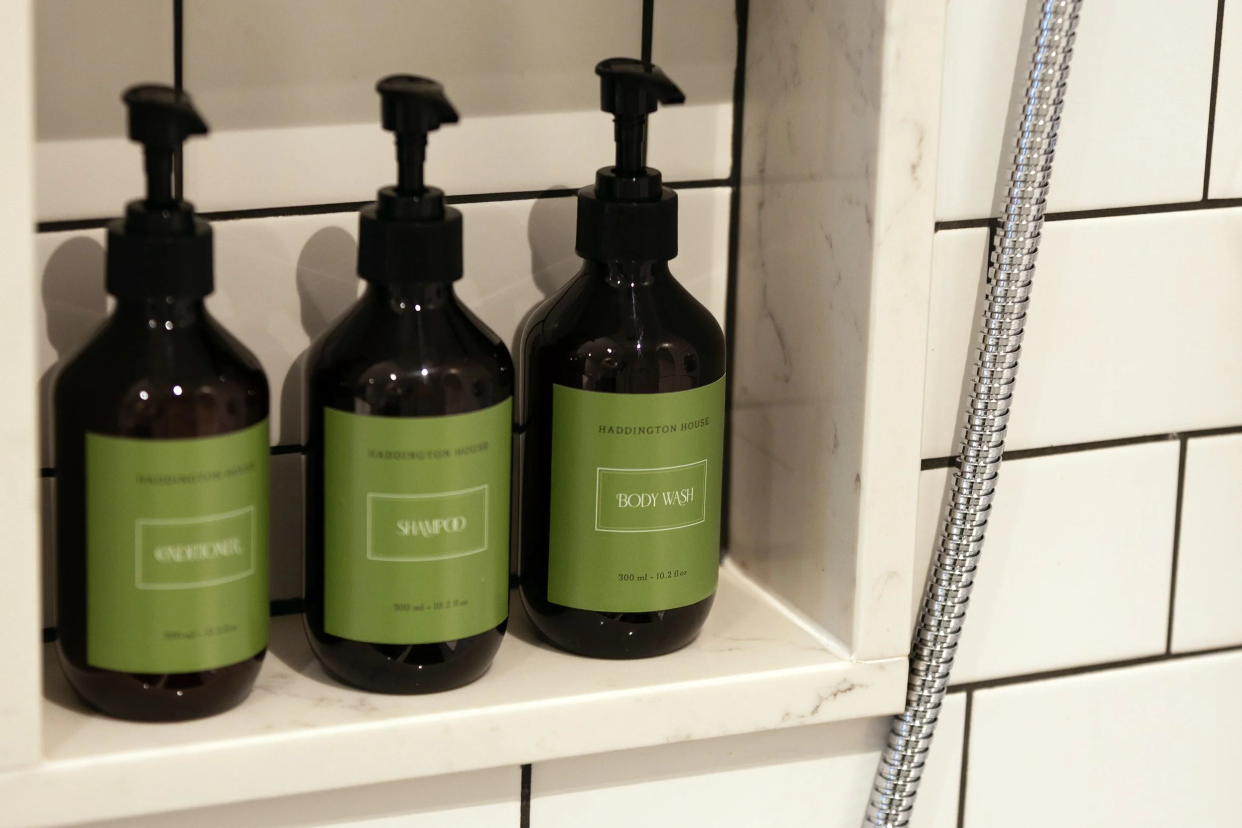 Three brown bottles of personal care products with green labels, placed on a white tiled shelf in a bathroom, next to a shower hose.