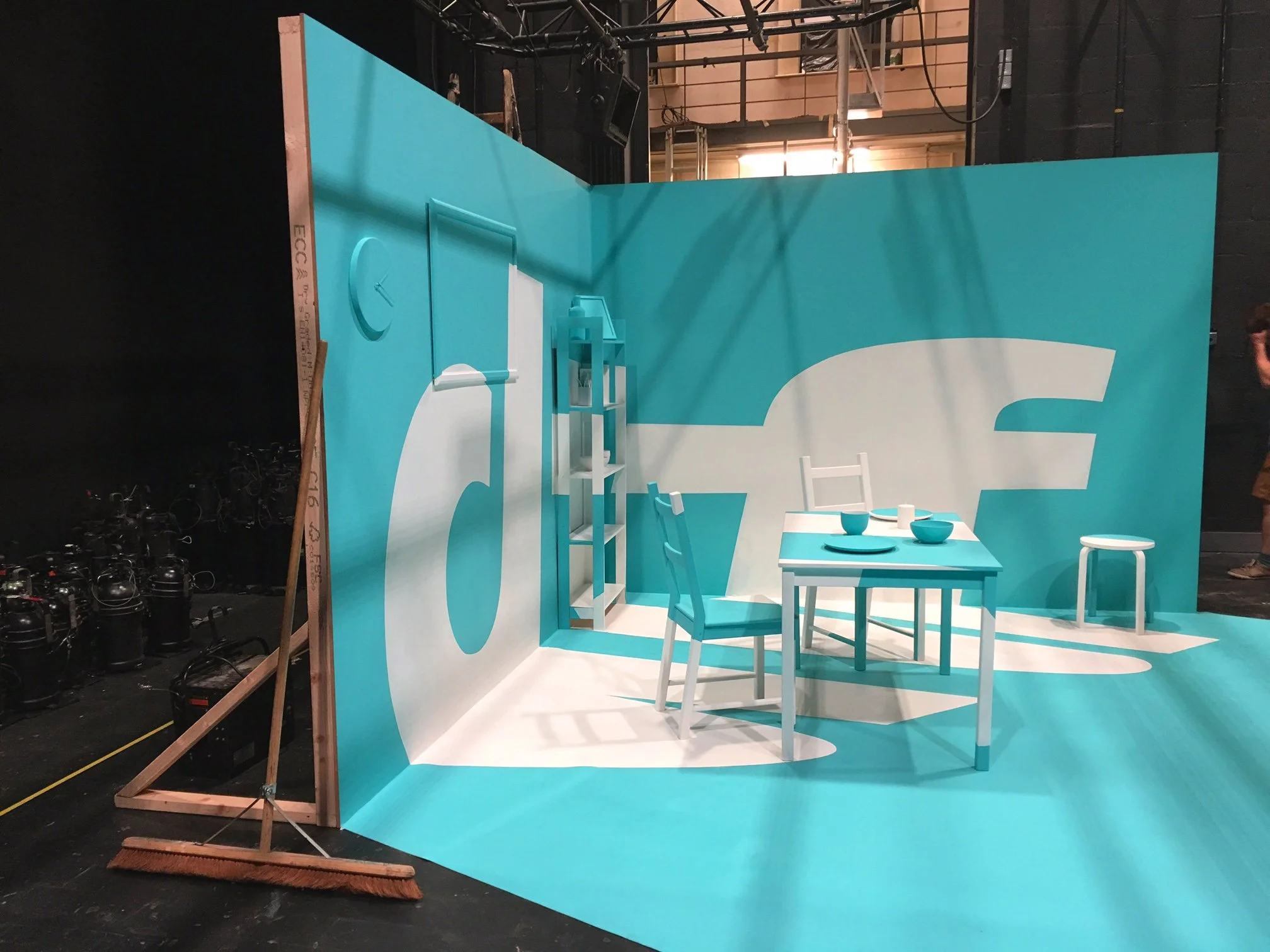 The image shows a blue-colored room with minimalist furniture, including a table, chairs, and shelves, inside a large studio or warehouse space with visible lighting and equipment.