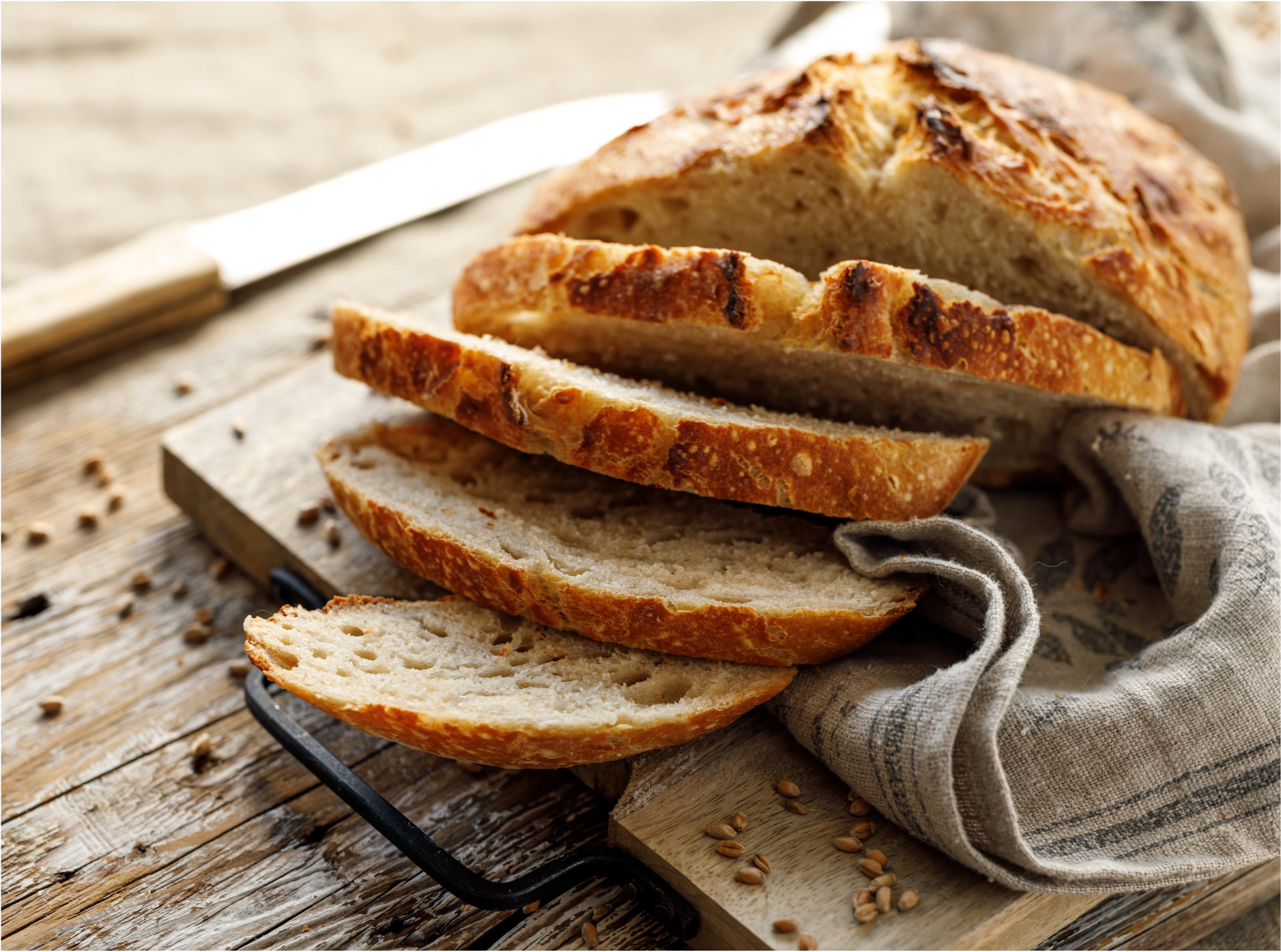 Sliced artisan bread on a wooden cutting board with a bread knife in the background and scattered grains around.