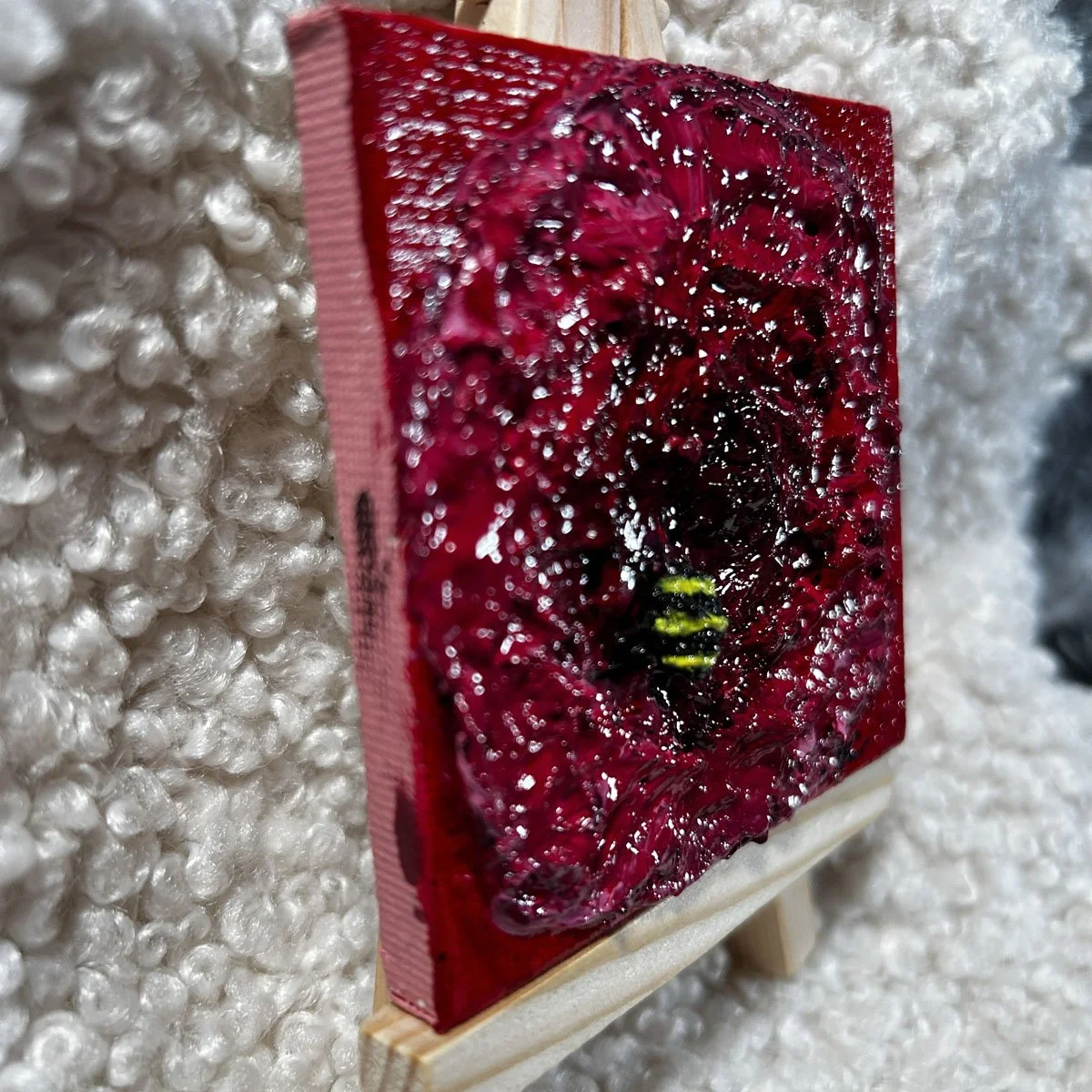 Side view of a small textured painting of a red flower with a raised bumblebee detail, showing thick layered paint and a glossy surface.