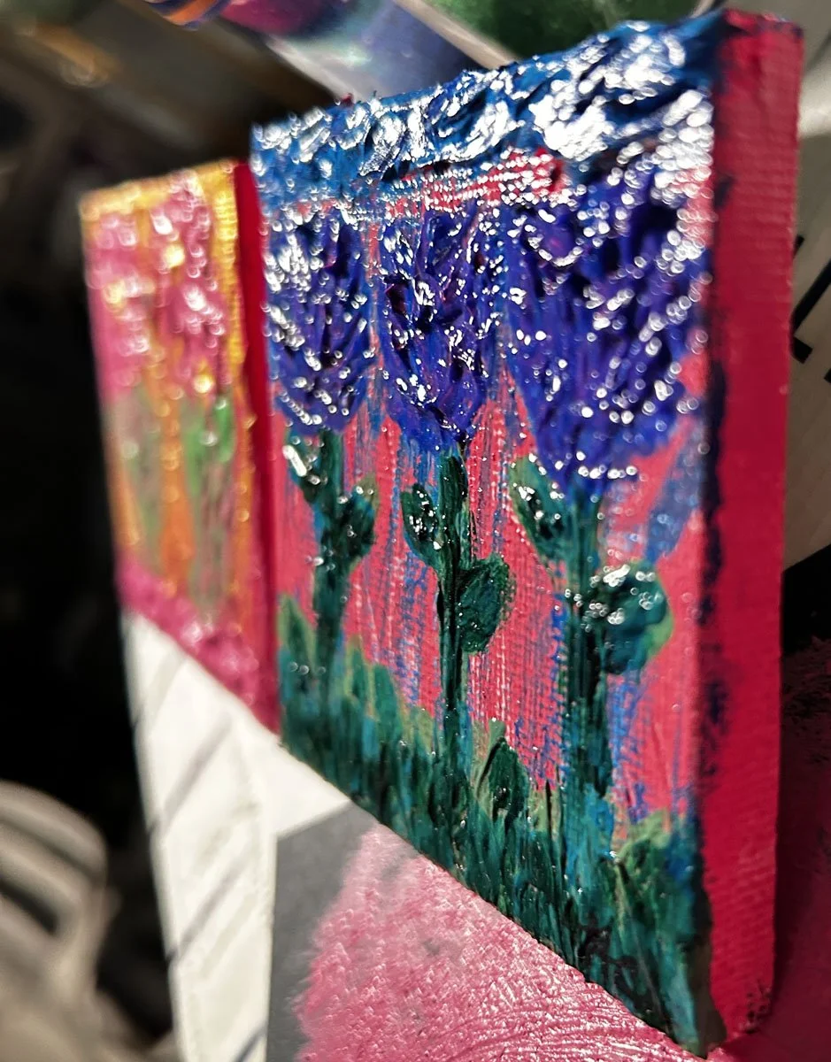 Side view of a small textured painting of purple flowers on a bright pink background, showing thick, glossy paint details on a mini canvas by Heather Collis