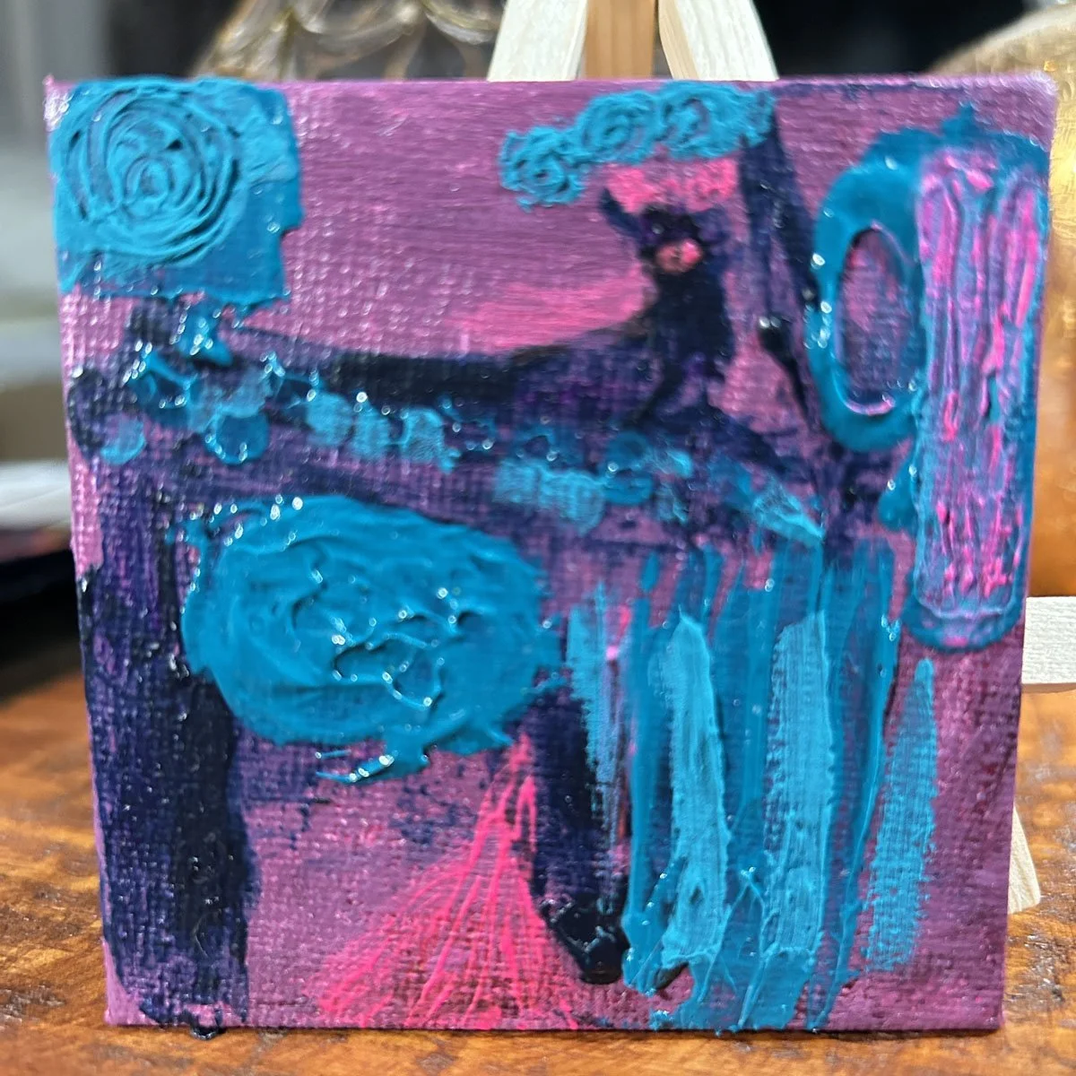 Close-up of a small square canvas with vibrant turquoise and dark blue impasto textures on a purple and pink background.
