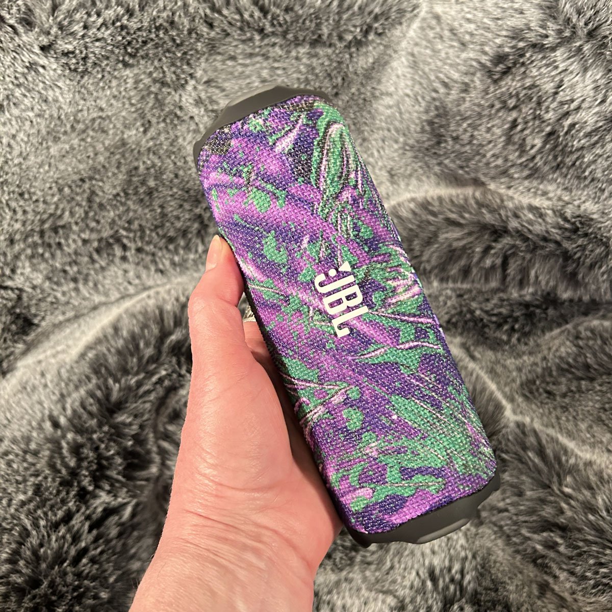 A hand holding a JBL portable Bluetooth speaker wrapped in a custom tech skin pattern showcasing a vibrant purple and green abstract painting design against a gray faux fur background.
