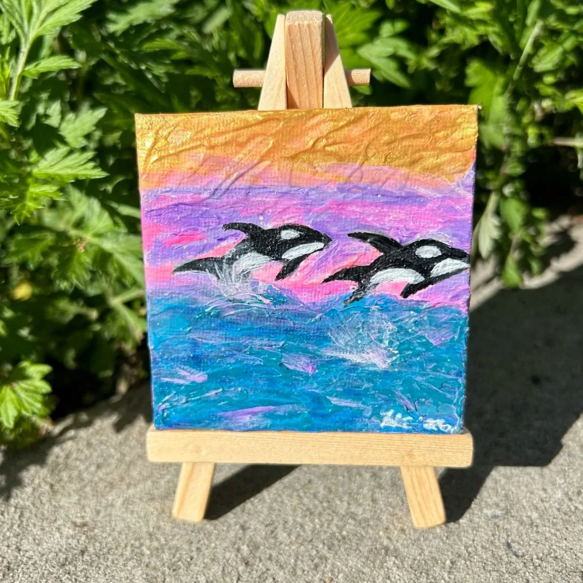 Killer Whales swimming in the ocean in a mini art painting on an easel with pink gold and purple by Heather Collis