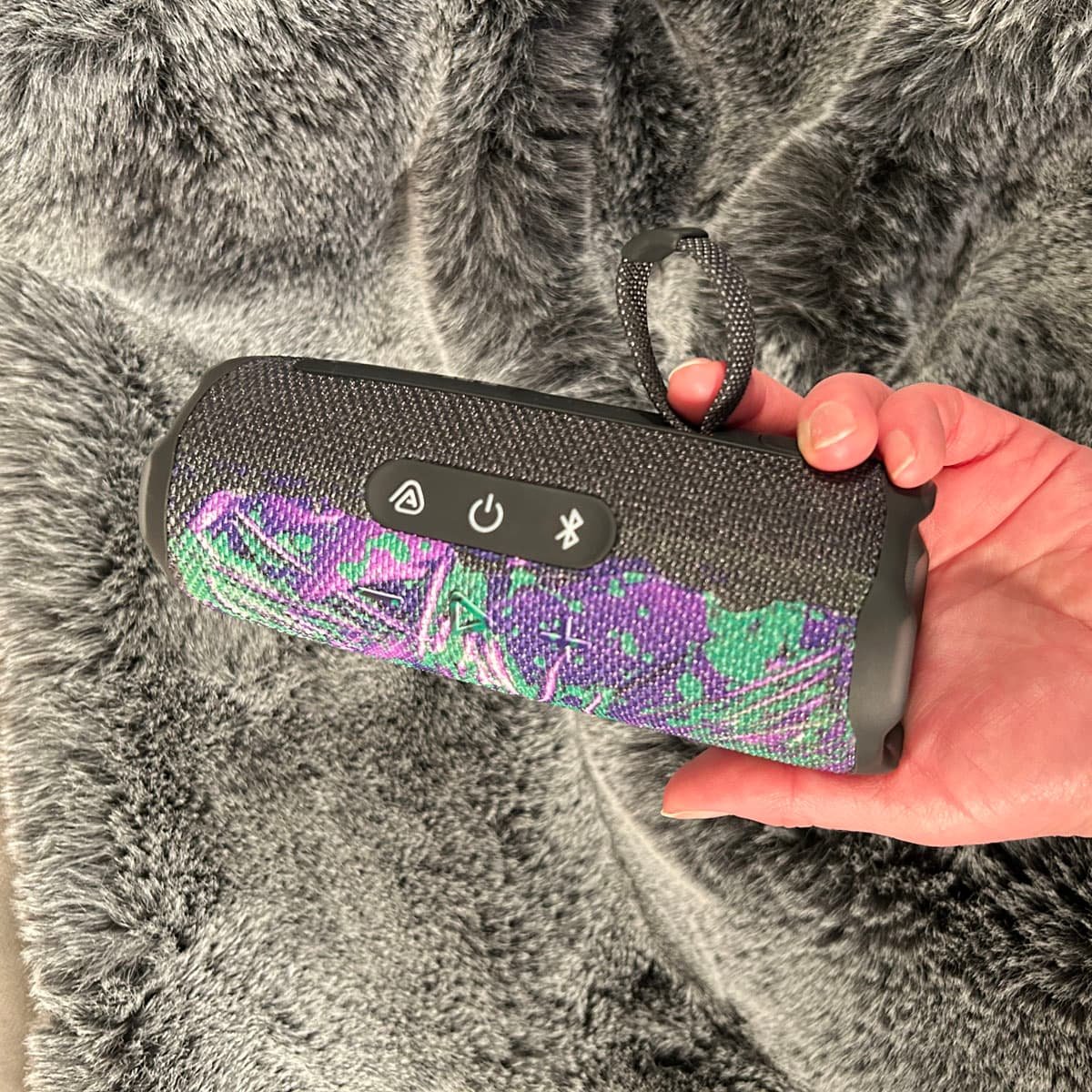 A vibrant purple and green abstract acrylic design pattern by Heather Collis Design wraps around a portable Bluetooth speaker, highlighting how the custom tech skin covers the device controls.
