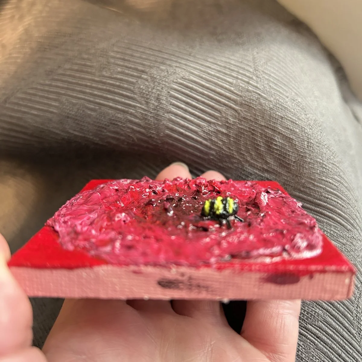 Side view of a small textured painting of a red flower with a raised bumblebee detail, showing thick layered paint and a glossy surface.