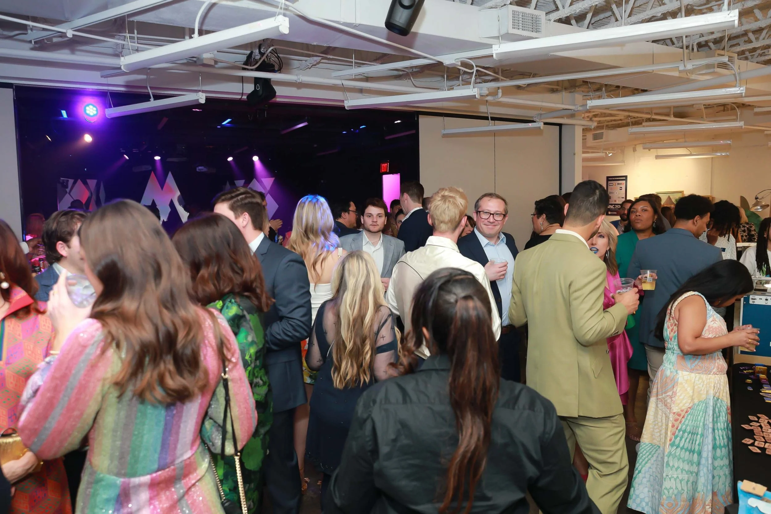 A crowded indoor event with diverse people mingling, some holding drinks, in front of a dark stage with purple lighting.