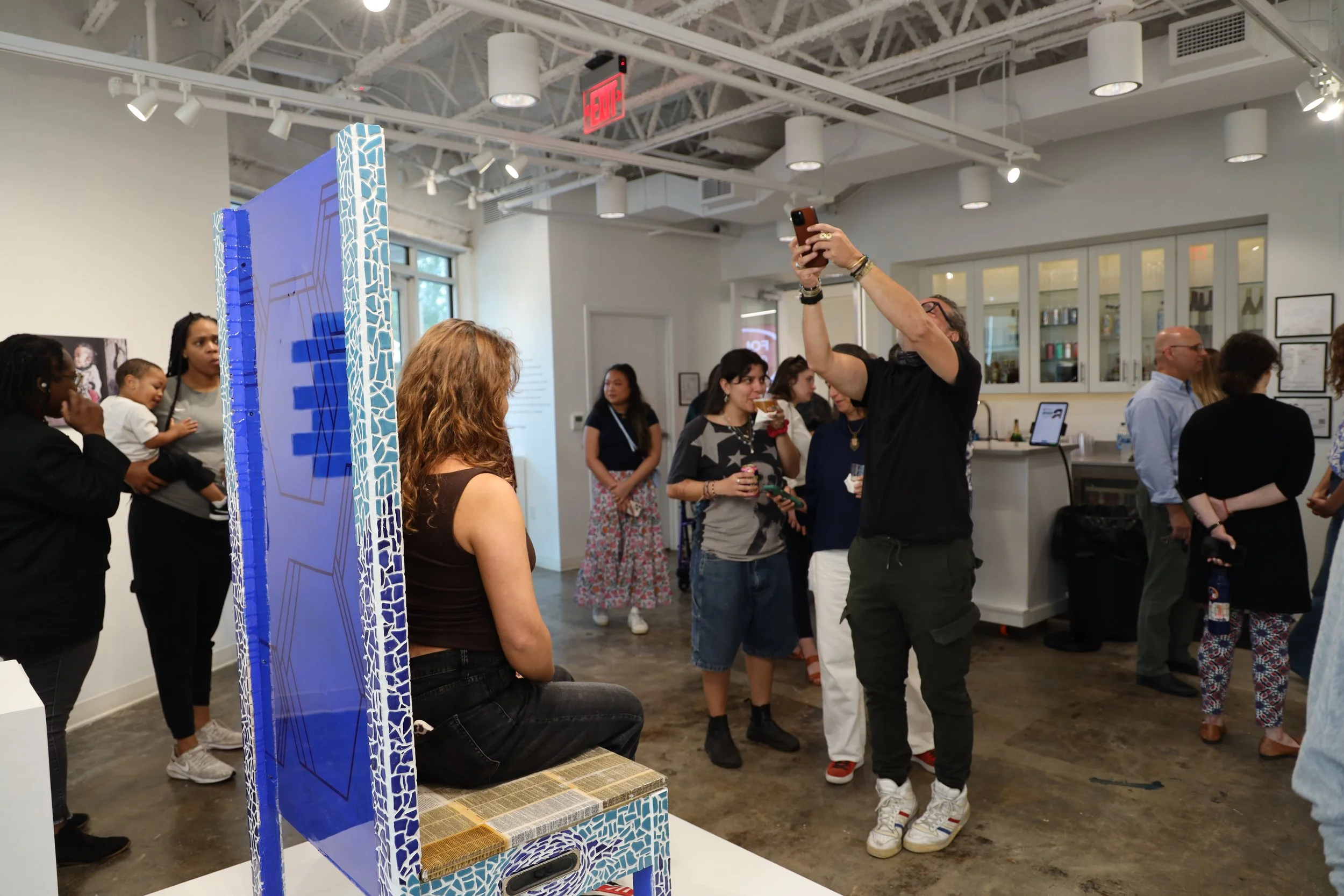 People gathered in an art gallery, some interacting with artwork, with one person taking a photo. The gallery interior features white walls, large windows, and display cabinets. There is a blue mosaic sculpture or partition in the foreground.
