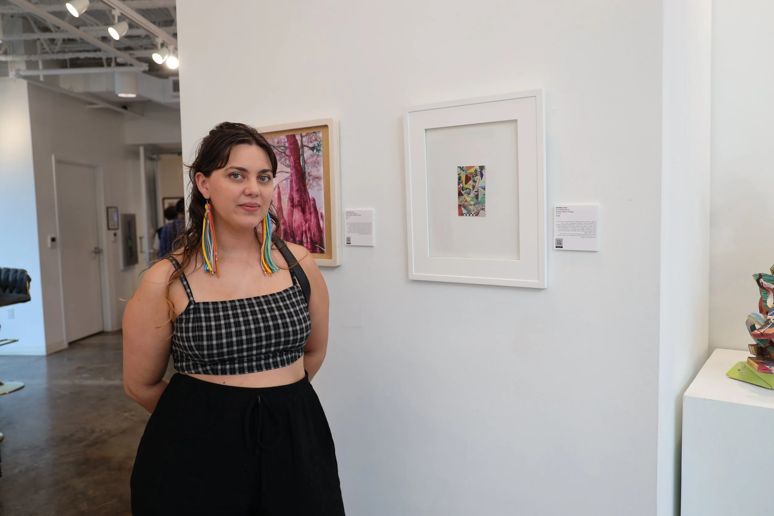 A woman with curly hair wearing a black and white checkered top and colorful earrings stands in an art gallery next to framed artwork on a white wall.